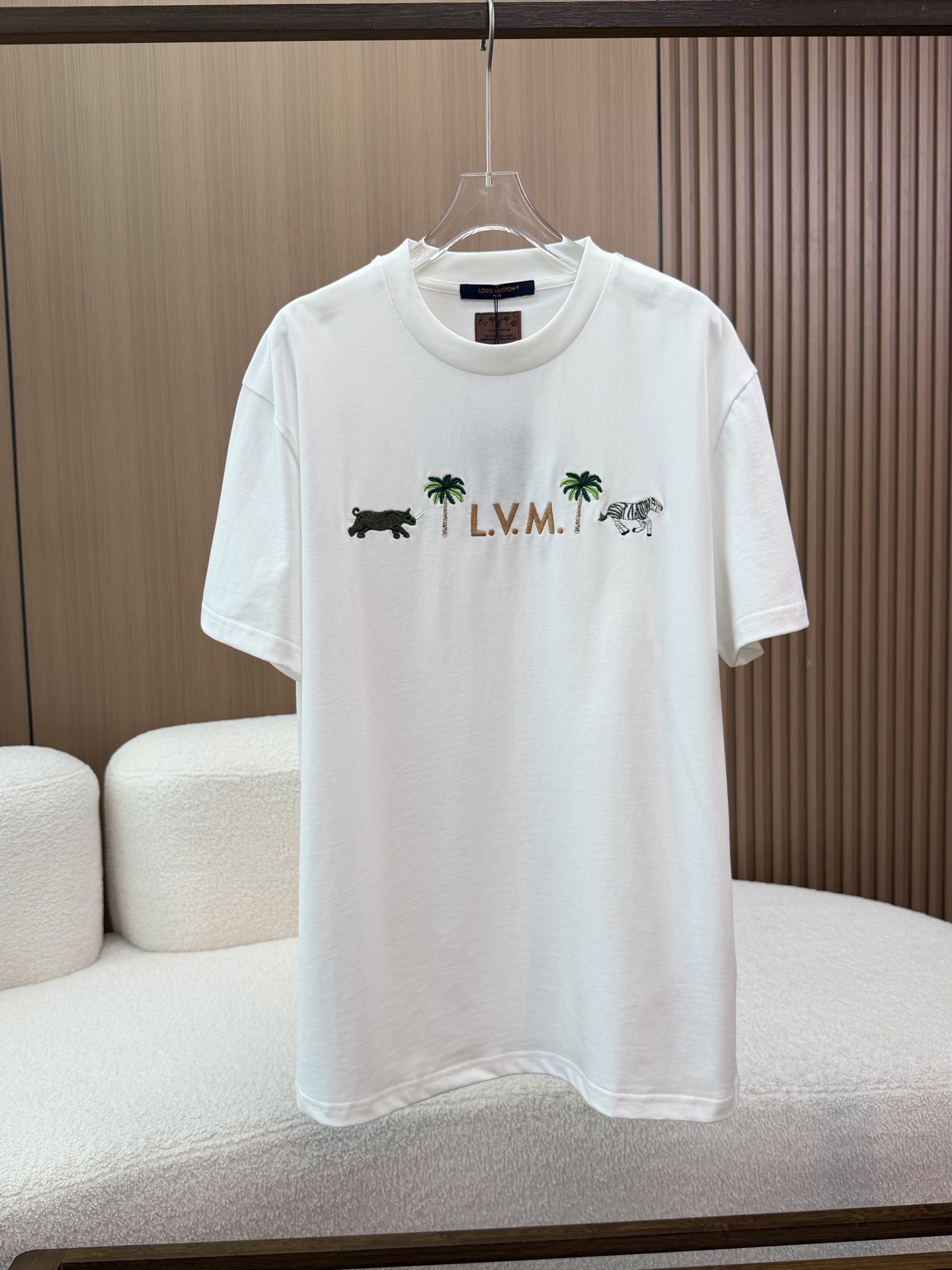 NO:863407,LV/Louis Vuitton 26ss new milky white cotton 3D letter palm animal embroidery short-sleeved T-shirt. This Louis Vuitton short-sleeved T-shirt is made of milky white cotton fabric, which is soft and skin-friendly, breathable and comfortable, suitable for daily wear. It has a classic round neck design, and the ribbed neckline is neatly routed, not easy to deform, and fits the neck line.  The front adopts exquisite embroidery technology, with a three-dimensional and full 'L.V.M.' letter embroidery in the center, palm tree patterns symmetrically distributed on both sides, a dynamic rhinoceros embroidery on the left, and a zebra embroidery on the right. The colors are layered and the stitching is delicate, highlighting the tropical style and brand recognition. A square leather label is sewn on the back of the neck with 'MARQUE L.VUITTON' printed on it.  DEPOSEE' words are outlined with reddish-brown stitching on the edge, which enhances the overall texture and recognition. The cuffs and hem adopt double-needle hemming technology, with neat routing and exquisite details, ensuring the durability and crispness of the clothing. , louis vuitton, louis vuitton, T-shirt, tees, t-shirt,19860909LV/路易威登 26ss新品乳白棉3D字母棕榈动物刺绣短袖T恤 这款路易威登短袖T恤采用乳白全棉面料,触感柔软亲肤,透气舒适,适合日常穿搭 经典圆领设计,罗纹领口走线工整,不易变形,贴合颈部线条. 正面采用精致刺绣工艺,中央是立体饱满的“L.V.M.”字母刺绣,两侧对称分布棕榈树图案,左侧为动感的犀牛刺绣,右侧为斑马刺绣,色彩层次分明,针脚细腻,彰显热带风情与品牌辨识度 后颈处缝制了一块方形皮革标签,印有“MARQUE L.VUITTON DEPOSEE”字样,边缘以红棕色缝线勾勒,提升了整体质感与辨识度 袖口与下摆采用双针锁边工艺,走线工整,细节考究,保证了衣物的耐穿性与挺括感,,louis vuitton,louis vuitton,T-shirt,tees，t-shirt,,Women's clothing