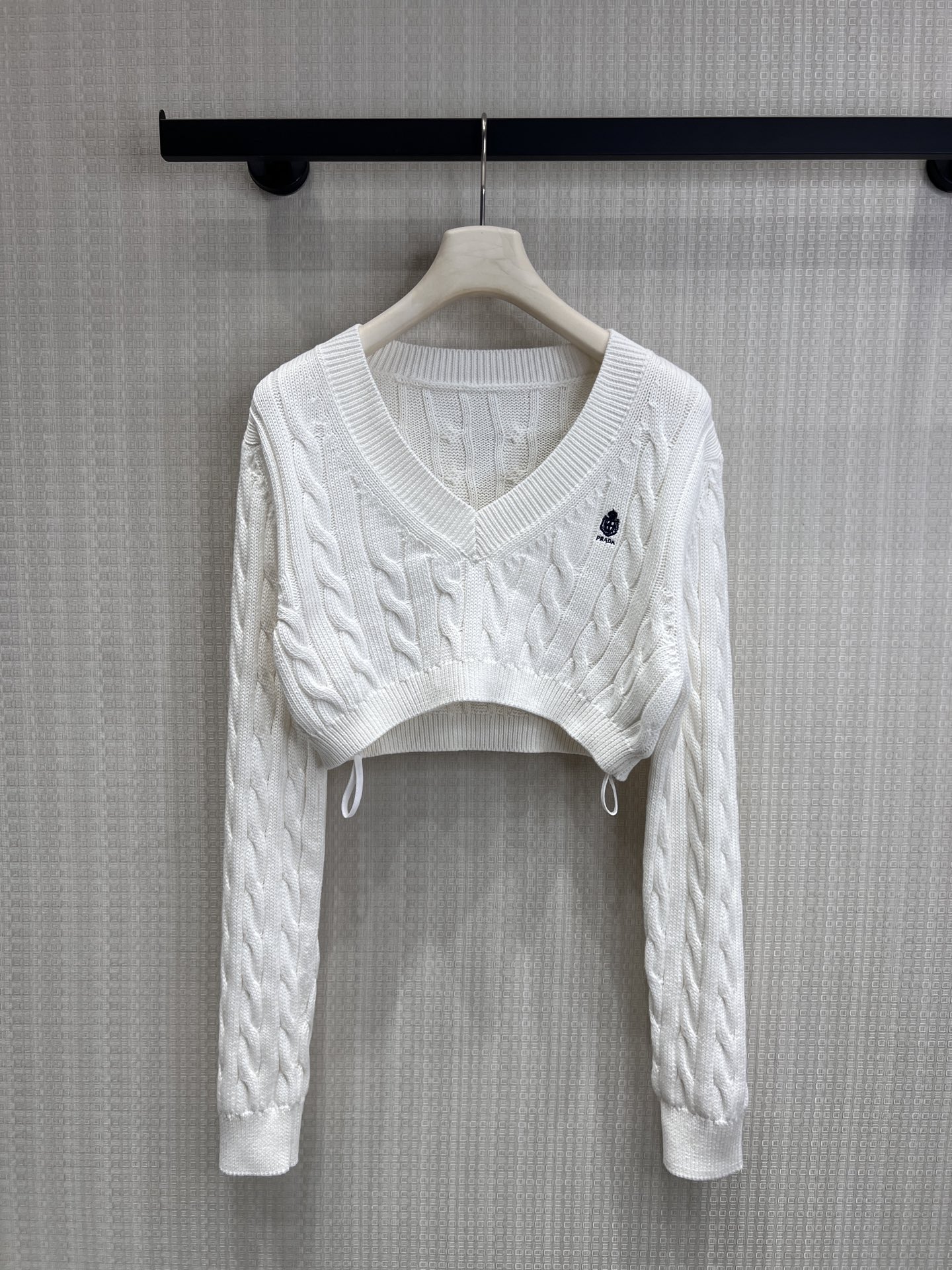 NO:844010,26 spring and summer new products  Pra**'s iconic college aesthetic is perfectly interpreted by this short cable knit sweater. The high-count cotton and linen blended yarn creates a full-textured twist texture, which is soft and skin-friendly. It is paired with a neat short cut to easily raise the waistline. The V-neck line neatly modifies the collarbone, and the embroidered logo on the chest low-key highlights the brand recognition.  The adjustable drawstring design at the hem is suitable for a variety of styles. The pure white color has a cool and noble air. Wearing it alone is the standard for Korean drama heroines in college. Pair it with a suit or denim items to instantly transform into a retro British style. It is easy to create a relaxed and refined atmosphere. Ready for sale smL, prada, business  suit,1986090926春夏新品 Pra**标志性学院美学,被这件短款绞花针织衫完美诠释.高支棉麻混纺纱线织出肌理饱满的麻花纹路,软糯亲肤的触感,搭配利落的短款剪裁,轻松拉高腰线,V领线条利落修饰锁骨,胸前刺绣徽标低调彰显品牌辨识别度,下摆可调节抽绳设计,适配多种穿搭格.纯净白调自带清冷贵气,单穿是韩剧女主的学院标配,搭配西装或牛仔单品,秒变复古英伦风,轻松拿捏松弛又精致的氛围感.现货出售smL,,prada,business suit,,Women's clothing