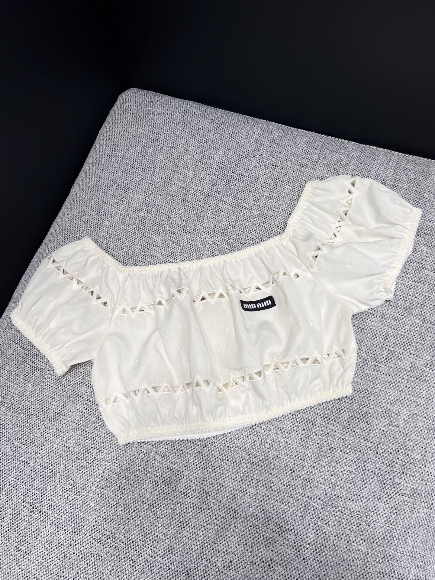 NO:893074,26 spring and summer new products  Miu** milk white hollow one-shoulder short top, replica of Italian retro sweet temperament. Soft waxy cotton material with soft matte texture, elastic one-line collar with short puff sleeves, gently showing off the shoulder and neck lines. The triangular hollow cut all over the body is transparent and light, the layers are exquisite but not exaggerated, and the mini woven label on the chest is low-key engraved  The brand logo neutralizes sweetness and adds light luxury. The short waisted version raises the waistline and is suitable for wearing as a complete set of hollow skirts in the same series. It is suitable for daily appointments and short trips. It is a gentle and smart gold-style top. In stock 36/38/40,, miumiu, skirts, T-shirt,1986090926春夏新品 Miu**奶白镂空一字肩短上衣,复刻意式复古清甜气质.软糯肌理棉料自带柔和雾面质感,松紧一字领搭配泡泡短袖,温柔展露肩颈线条.周身错落三角镂空剪裁通透轻盈,层次精致不浮夸,胸前mini织标低调镌刻品牌标识,中和甜腻增添轻奢底蕴.短款收腰版型拉高腰线,适配同系列镂空半裙成套穿搭,日常约会、短途出游皆适配,温柔又灵动的千金风上衣.现货36/38/40,,miumiu,skirts,T-shirt,,Women's clothing