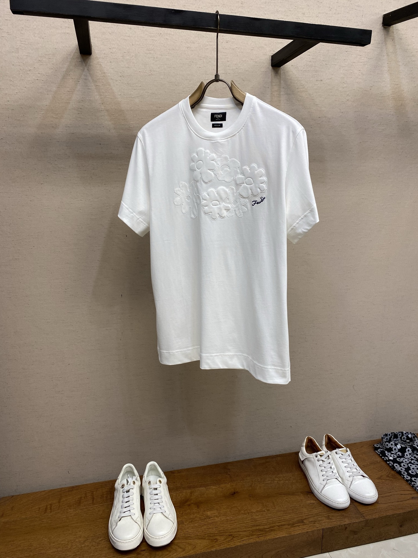 NO:834437,Fendi, appliqué embroidered round neck short-sleeved T-shirt fabric selection  It is made of pure cotton material with a weight of 220g. It is derived from natural cotton fiber. It has a soft and delicate touch, is skin-friendly and non-irritating, and can be worn by sensitive skin with peace of mind. Using the 20S/1 compact spinning process, the yarn has less hairiness, even dryness, clear and regular texture, and is visually more sophisticated. At the same time, it retains the natural breathable holes of cotton fiber and has excellent moisture absorption and perspiration effects. Compared with ordinary plain cotton, the compact cotton cloth is more wear-resistant, less prone to pilling, and more durable for daily wear. Back neck embroidery  The same color high-gloss embroidery thread is used, and the embroidery machine works with high precision. The stitches are closely arranged without skipping or missing stitches. The edges of the logo are clear and sharp, showing a three-dimensional and full visual effect. You can feel the uniform raised texture when touching, solid and layered; the embroidery area is accurately located in the center of the lower back collar, with coordinated proportions and no offset, skew and other problems. Blue letter embroidery  The flowers are embroidered with blue polyester fiber embroidery thread, forming a soft contrast with the white background. The handwritten embroidery restores the original brushstroke details, with smooth and natural lines; the embroidery stitches are fine and firm, and will not easily come off or turn white after being worn for a long time. Appliqué Embroidery  The flowers are made of pure cotton patches of the same color. The patch fabric has the same origin as the main fabric of the T-shirt, and the texture is highly unified, which avoids the abrupt feeling of splicing different materials. The edges of the patch have been overlocked to prevent curling and threading. At the same time, the same-color design makes the flower pattern low-key and layered, showing subtle texture changes under the light. Details  1. The neckline is made of 285g/㎡ high-elastic ribbing, which has enough stiffness to support without losing the soft and skin-friendly touch. The thickness is 50% thicker than the body fabric. It will not deform or collapse after repeated pulling, and it fits the neck without being tight. 2. After pre-shrunk treatment, the shrinkage rate is less than 2, ensuring that the shape can be kept stiff after washing, and the embroidery will not be tight or wrinkled.  Size S-XL,,fendi,T-shirt,tees,t-shirt,19860909Fendi,贴布绣花圆领短袖T恤 面料选择 选用纯棉材质,克重达220g,源自天然棉纤维,触感柔软细腻,亲肤不刺激,敏感肌也能安心穿着.采用20S/1紧密纺工艺,纱线毛羽少、条干均匀,纹理清晰规整,视觉上更显精致高级.同时保留了棉纤维的天然透气孔,吸湿排汗效果出色相比普通平纹棉,紧密棉的布面更耐磨、不易起球,日常穿着更耐造. 后领刺绣 选用同色系高光泽绣线,通过绣花机高精度作业,针脚紧密排列无跳线、漏针现象,标志边缘清晰锐利,呈现出立体饱满的视觉效果,触摸时能感受到均匀的凸起质感,扎实且富有层次；刺绣区域精准位于后领下方中央,比例协调,无偏移、歪斜等问题. 蓝色字母刺绣 花朵旁饰以蓝色聚酯纤维绣线刺绣,与白色底色形成柔和撞色,手写体的刺绣还原了原版的笔触细节,线条流畅自然；刺绣针脚细密牢固,久穿不易脱线、不易发白. 贴布绣 花朵采用同色系纯棉贴布打造,贴布面料与T恤主体面料同源,质感高度统一,避免了不同材质拼接的突兀感；贴布边缘经过锁边处理,不易卷边、不易脱线,同时同色系设计让花朵图案低调又有层次,在光线下能呈现出微妙的肌理变化. 细节 1领口采用285g/㎡高弹罗纹,拥有足够的挺括感来支撑,又不失柔软亲肤的触感,厚度比衣身面料厚50,反复拉扯也不会变形、垮塌,同时贴合颈部却不紧绷. 2通过预缩水处理之后缩水率低于2,保证洗涤之后能保持版型挺括,绣花处不会紧绷起皱. 尺码S-XL,,fendi,T-shirt,tees，t-shirt,,Men's clothing
