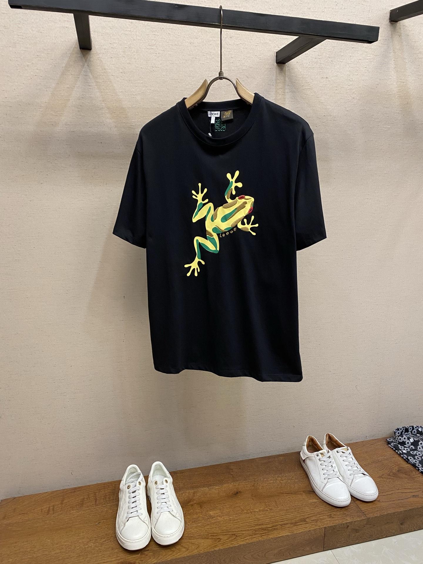 NO:834460,Loewe, frog print embroidered crew neck short sleeves. Original purchase; pure cotton knitted fabric, decorated with embroidery and frog print pattern on the front, Anagram embroidery on the back, loose fit, standard length, round neck design, ribbed collar, full set of accessories # Made of pure cotton knitted fabric Made of 24-count special-spun compact cotton, 185g/㎡; paired with 32-count 1×1 rib, 340g/㎡. It is a medium-thick knitted fabric that is both stiff and comfortable. In the finishing stage before embroidery, the fabric is tentered and set by a high-temperature setting machine, and the tension is The control makes the warp and weft yarns return to straightness and width, eliminating wrinkles and internal stress caused by the previous process. The compact spinning technology itself has made the fibers arranged parallel, hairiness significantly reduced, and the surface smooth and smooth; the fabric after shaping has both stiffness and softness, and a moderate drape. # The front is decorated with an embroidered frog print pattern. The front frog pattern is printed with environmentally friendly elastic glue and then embellished with positioning needle embroidery. The combination of the two processes makes the pattern have the full color block of glue printing and the three-dimensional texture of embroidery. The background color of the frog is printed on the surface of the fabric through environmentally friendly elastic glue. The glue has excellent elasticity and adhesion, can extend synchronously with the stretching of the fabric, is not easy to crack and fall off, and complies with environmental standards. The frog's eyes, patterns and other details are embellished and embroidered using the positioning needle embroidery process. The needle embroidery is a stitching method in which short stitches are closely arranged. The stitches are flat and the edges are sharp, which can accurately present the delicate details of the frog pattern. # Anagram embroidery on the back Made with three-dimensional embroidery technology, three-dimensional embroidery is an embroidery technique that uses special stitches to make the pattern appear in relief by superimposing embroidery threads and filling pads. The three-dimensional embroidery has a higher stitch density and richer layers, which can form obvious concave and convex touches and light and shadow changes. The high-density stitch stacking creates a raised effect; after the embroidery is completed, the padding is removed by washing, leaving a full three-dimensional logo pattern. # Standard round neck design This short-sleeve style is designed with a loose fit and a standard length. The shoulder line slightly exceeds the acromion point, showing a slight shoulder drop effect; the body is straight cut, and the bust and hem circumferences are similar, creating a relaxed and comfortable wearing feeling. The standard length avoids the feeling of being too long and makes the overall proportion more coordinated. Sizes XS-L,, loewe, T-shirt,19860909Loewe,青蛙印绣圆领短袖 原购买；纯棉质针织面料、正面饰有刺绣和青蛙印花图案、背面Anagram刺绣、宽松版型、标准长度、圆领设计、罗纹衣领、全套辅料配备 # 纯棉质针织面料材质 采用24支特纺紧密棉,185g/㎡；搭配32支1×1罗纹,340g/㎡.属于兼具挺括与舒适的中厚型针织面料.在刺绣前的整理阶段,面料经高温定型机进行拉幅定形,通过张力控制使经纬纱恢复平直、门幅一致,消除前道工序产生的褶皱与内应力.紧密纺技术本身已使纤维排列平行、毛羽显著减少,表面光洁平滑；定形后的面料兼具挺括性与柔软度,垂坠感适中. # 正面饰有刺绣青蛙印花图案 正面青蛙图案采用环保弹力胶浆印花打底后,再以定位挨针刺绣进行点缀的复合工艺.两种工艺结合,使图案既有胶浆印花的饱满色块,又有刺绣的立体质感. 青蛙的底色通过环保弹力胶浆印于面料表面,该胶浆具有优异的弹性与附着力,能随面料拉伸而同步延展,不易开裂脱落,同时符合环保标准. 采用定位挨针刺绣工艺对青蛙的眼睛、花纹等细节进行点缀刺绣.挨针刺绣是一种短针脚紧密排列的针法,针迹平整、边缘锐利,能精准呈现青蛙图案的精致细节. # 背面Anagram刺绣 采用立体刺绣工艺制作,立体刺绣是一种通过叠加绣线、填充衬垫.使用特殊针法使图案呈现浮雕效果的刺绣技法,立体刺绣的针迹密度更高、层次更丰富,能形成明显的凹凸触感与光影变化.通过高密度针脚堆叠形成隆起效果；刺绣完成后通过水洗去除衬垫,留下饱满立体的标志图案. # 标准圆领版型设计 这款短袖采用宽松版型搭配标准长度设计.肩线略微超过肩峰点,呈现轻微落肩效果；衣身呈直筒剪裁,胸围与下摆围度相近,营造出松弛从容的穿着感.标准长度避免了过长的拖沓感,使整体比例更加协调. 尺码XS-L,,loewe,T-shirt,,Men's clothing