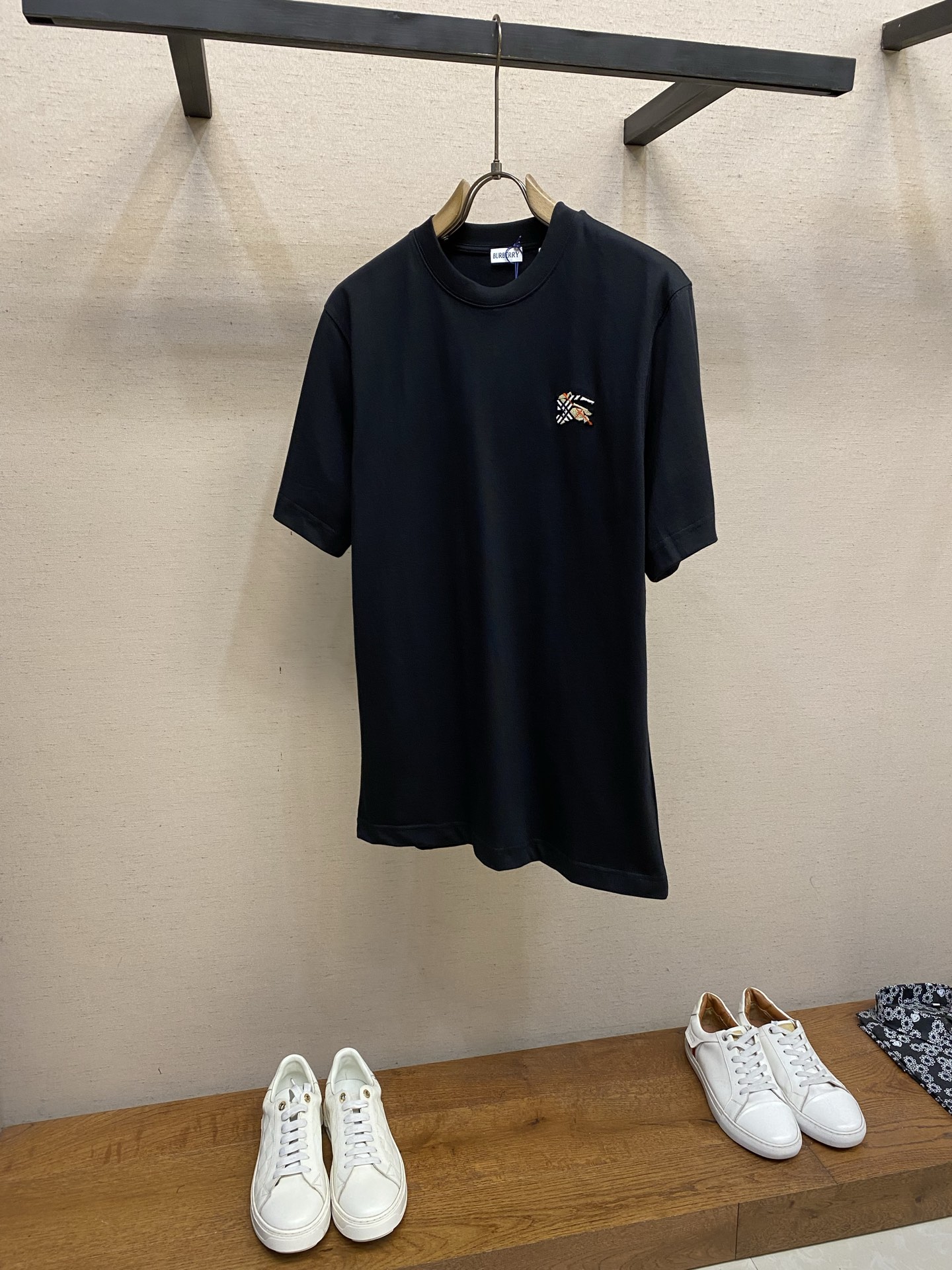 NO:838914,P BBR, plaid toothbrush embroidered round neck short sleeves. Available in black, white, green. Original purchase; light plain woven pure cotton fabric, embellished with tufted equestrian knight logo embroidery, integrated with the iconic sand beige plaid, and a full set of accessories. # Lightweight plain woven compact cotton fabric  Use 15-count special-spun compact cotton, 230g/㎡; match 32-count 1×1 rib, 340g/㎡. The fabric is a heavy-weight fabric with thick needles, strong texture, and crisp texture. Because the 15-count yarn is thicker, the shaping process is particularly important, and it can  It eliminates the internal stress generated during the weaving process of thick yarns and prevents pattern misalignment due to fabric shrinkage during subsequent multi-layer printing. The collar is a high-density tight rib made of cotton, which achieves elasticity through high-density weaving, making the touch more pure and natural.  # Embellished tufted equestrian knight logo embroidery The equestrian knight logo uses tufted three-dimensional toothbrush embroidery technology. This technology is characterized by dense and straight piles, soft to the touch like velvet, and clear edge outlines.  This is an embroidery technique that uses special needles to embed embroidery threads into bundles into the base fabric to create an upright velvet effect. The embroidery needle pierces the fabric with high-speed reciprocating motion, bringing in multiple strands of embroidery thread at the same time and forming a coil; after the embroidery is completed, the back is coated with water-based adhesive to fix the bottom of the coil, and the front is cut and trimmed to a uniform height through a shearing process to form an upright texture similar to toothbrush bristle bundles. # Standard casual style  It adopts a casual version design, which is between the standard version and the loose version. The core feature is that the shoulder line slightly exceeds the acromion point, showing a slight shoulder drop effect; the bust looseness is about 12-16cm compared to the net size, and the body is straight cut; the sleeve shape is wider than standard short sleeves, and the sleeve length is slightly longer. Matched with 230g thick needle weight fabric  When matched, the stiffness of the fabric supports the structure of the casual version - it is neither too loose like the loose version, nor as rigid as the standard version. The advantage of the casual version is that it is 'relaxed and moderate', complementing the classic British temperament of the equestrian knight logo, and is suitable for switching between daily leisure and sports scenes.  Size XS-L,,T-shirt,19860909P BBR,格纹牙刷绣圆领短袖 发售色系黑色、白色、绿色 原购买；轻盈平织纯棉面料、点缀簇绒马术骑士徽标刺绣、融合标志性沙米色格纹、全套辅料配备 # 轻盈平织紧密棉面料 用15支特纺紧密棉,230g/㎡；搭配32支1×1罗纹,340g/㎡.面料属于粗针重克重面料,肌理感强、质地挺括.由于15支纱线较粗,定形工序尤为重要,它能消除粗支纱线织造过程中产生的内应力,防止后续多层印花时因面料收缩导致图案错位.衣领属于全棉高密度紧实罗纹,全棉罗纹通过高密度织造实现弹性,触感更加纯粹自然. # 点缀簇绒马术骑士徽标刺绣 马术骑士徽标采用簇绒立体牙刷刺绣工艺,该工艺的特点是绒毛密实挺拔、触感柔软如天鹅绒、边缘轮廓清晰. 这是一种通过特殊针具将绣线成束植入底布、形成竖立状绒毛效果的刺绣技法.刺绣针以高速往复运动刺穿面料,将多股绣线同时带入并形成线圈；刺绣完成后,背面涂覆水性背胶固定线圈底部,正面则通过剪毛工序将线圈顶部剪开并修剪至统一高度,形成类似牙刷毛束的直立质感. # 标准休闲版型 采用休闲版型设计,介于标准版与宽松版之间.核心特点为肩线略微超过肩峰点,呈现轻微落肩效果；胸围松量比净尺寸大约12-16cm,衣身呈直筒剪裁；袖型宽于标准短袖,袖长略长.与230g粗针重克重面料搭配时,面料的挺括性支撑了休闲版型的结构感——既不会像宽松版那样过度松垮,也不会像标准版那样拘谨.休闲版型的优势在于“松弛有度”,与马术骑士徽标的经典英伦气质相得益彰,适合日常休闲与运动场景的切换. 尺码XS-L,,T-shirt,,Men's clothing
