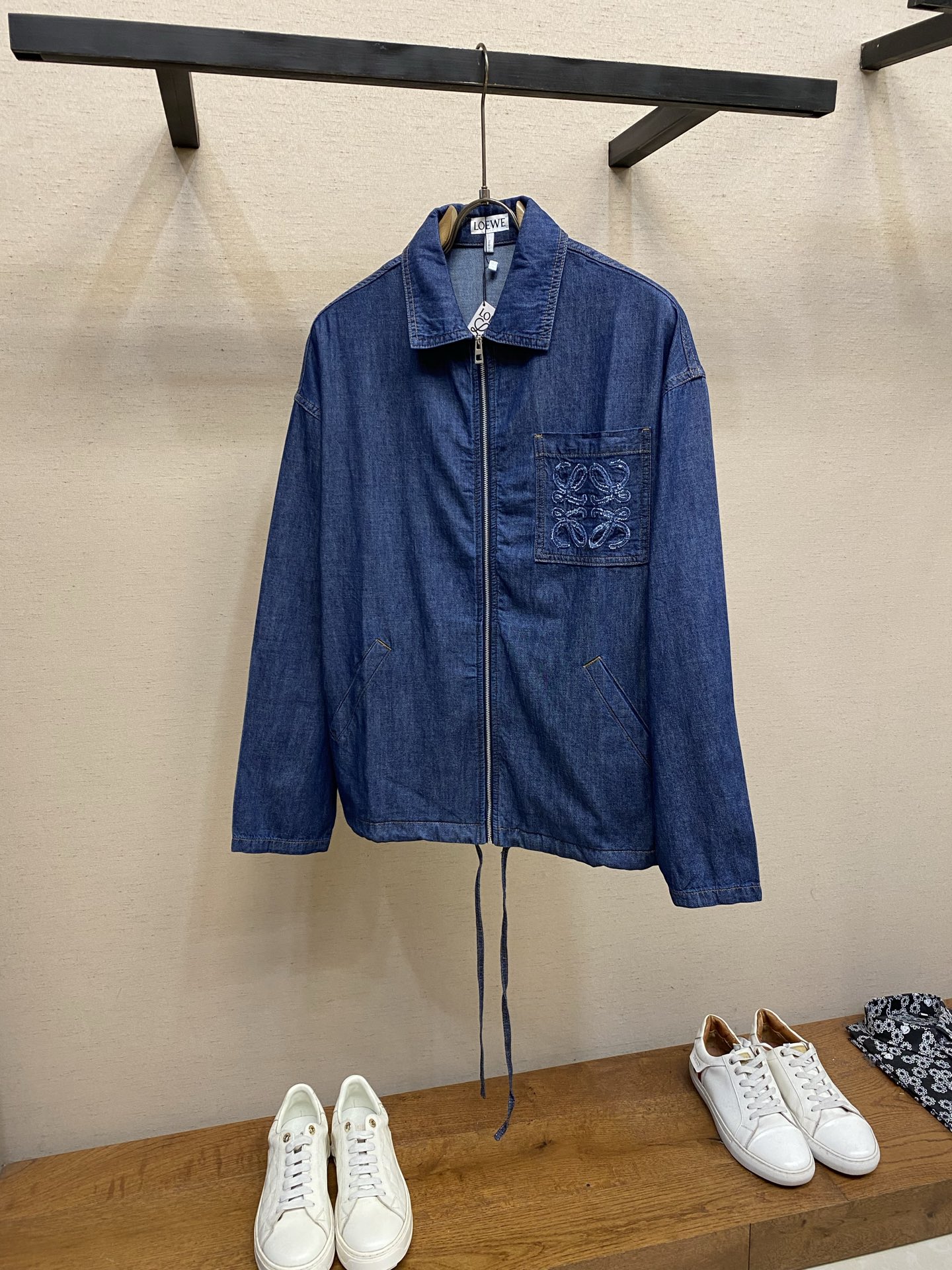 NO:947037,(Top), Loewe Patch Pocket Embroidered Denim Jacket. Fabric selection. Made of lightweight cotton chambray denim fabric, made with plain weave technology. The warp and weft cotton yarns are evenly interwoven, and the fabric surface presents a delicate and clear denim grain texture. It feels soft and thin, and it has a moderate drape. It is stiff and not easy to wrinkle and deform. The gray fabric is washed and pre-shrunk to remove the hard starch feeling of the fabric. The upper body is light and pressure-free, breathable, perspiration-wicking and not stuffy. Embroidery technology  The Anagram pattern is created using an exclusive appliqué process. First, the cut denim pattern pieces are accurately positioned on the patch bag base, and fixed on the positioning frame of the embroidery machine to prevent deviation. The needle is double-layered and densely embroidered to lock the fabric pieces along the outer contour of the pattern, and special special  The raw edge embroidery stitch method outlines the edge, and the embroidery threads are staggered and stacked to create a fluffy and three-dimensional tassel texture. The multi-layer lock embroidery structure firmly fixes the patch and is not easy to warp and peel off. The tassel texture is full and three-dimensional. After manual refinement, the lines are clean and neat, with both three-dimensional visual texture and solid firmness.  Details 1. Classic lapel structure, double-layer panels at the collar are overlocked and reinforced, and double open-stitched seams on the inside of the collar are not easy to roll and deform; 2. Embroidered patch pockets on the chest are matched with diagonal side pockets on both sides. All pockets are reinforced with double open-stitching, and the corners are tightened and locked to prevent cracking and deformation when objects are placed daily. The diagonal pockets fit the body, taking into account both beauty and practicality.  3. The hem has a built-in drawstring made of the same cotton material and is fixed with metal air holes, so the tightness of the hem can be adjusted freely; the cuffs are double-layered and hemmed with double-thread sewing, and the seams are dense and washable, so there will be no problems with thread breakage or fabric tearing. Sizes S-XL,, loewe, jackets & coats,19860909（上衣）,,Loewe 贴袋绣花牛仔外套  面料选择 选用轻盈棉质青年布牛仔面料,采用平纹紧密梭织工艺打造,经纬棉纱均匀交织,布面呈现细腻清晰的牛仔颗粒肌理,触感柔软轻薄,同时其自带适度垂坠感,挺括不易起皱变形.坯布经水洗预缩软化处理,去除布料硬性浆感,上身轻盈无压,透气排汗不闷肤.  绣花工艺 采用专属贴布绣工艺打造Anagram纹样,先将裁剪成型的牛仔布图案裁片精准对位铺置在贴袋基底上,固定于绣花机定位框防止走位偏移.针头沿图案外轮廓双层密绣锁固布片,同步运用特殊毛边绣针法勾勒边缘,绣线交错堆叠塑造蓬松立体的流苏肌理,多层锁绣结构牢牢固定贴布,不易翘边脱片,流苏纹理层次饱满立体,手工精修后线条干净利落,兼具立体视觉质感与扎实的牢固度.  细节 1、经典翻领结构,领部双层裁片锁边加固,领口内侧双道明线锁缝,不易翻卷变形； 2、胸前绣花贴袋搭配两侧斜插侧袋,所有袋口全部采用双道明线加固,转角位置收针锁结,日常放置物件拉扯不易开裂变形,斜插袋贴合衣身,兼顾美观与实用性. 3、衣摆内置棉质同料抽绳,搭配金属气眼固定,可自由调节下摆松紧；袖口双层折边双车线缝制,锁边密实耐洗,不会出现断线、布料撕裂问题. 尺码S-XL,,loewe,jackets & coats,,Men's clothing