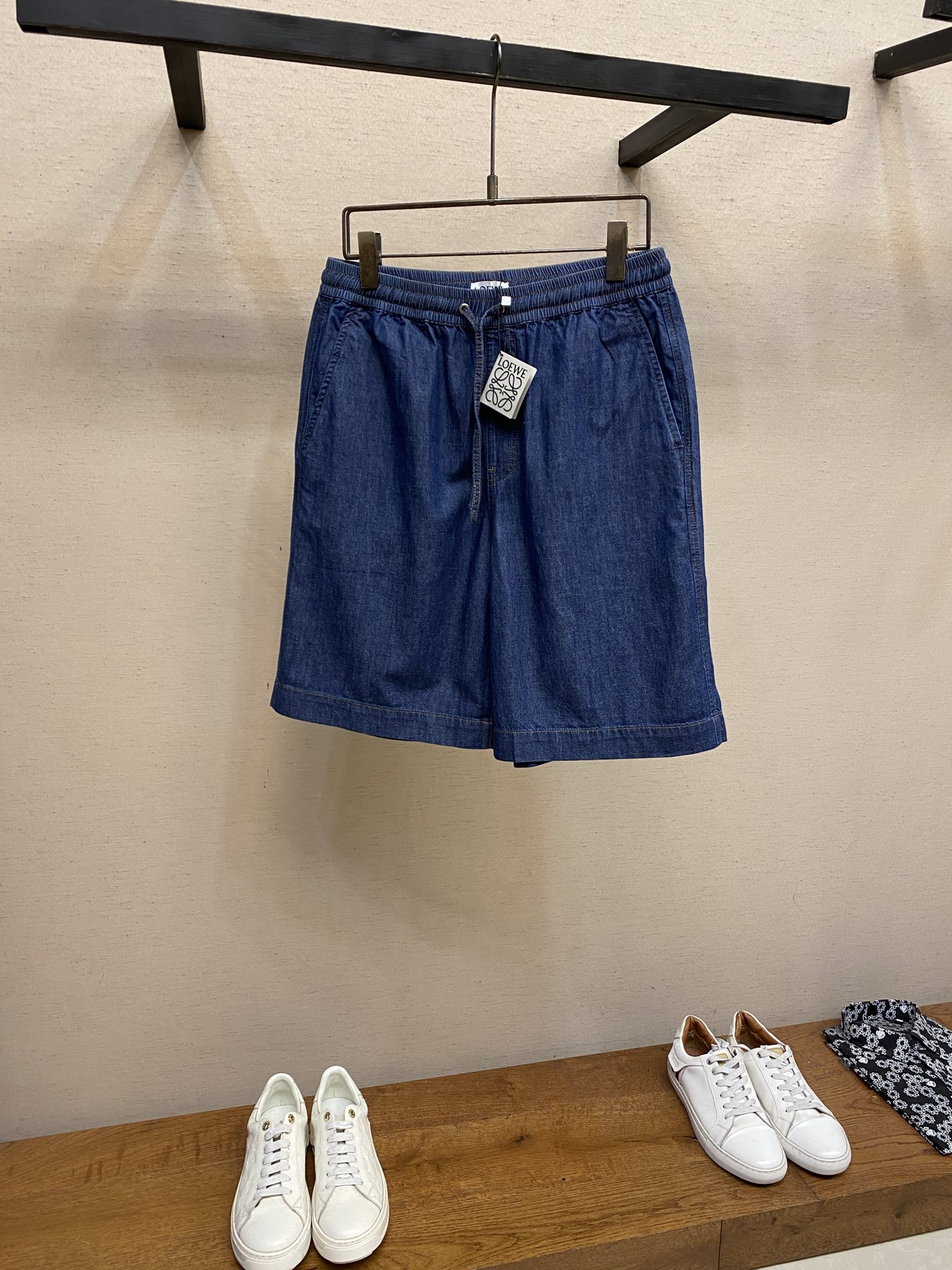 NO:947029,(shorts), Loewe Patch Pocket Embroidered Denim Shorts Fabric Selection: Made of lightweight cotton chambray denim fabric, made with plain weave and close weaving technology. The warp and weft cotton yarns are evenly interwoven, and the fabric surface presents a delicate and clear denim grain texture. It feels soft and thin, and has a moderate drape feeling. It is stiff and not easy to wrinkle and deform. The gray fabric is washed, pre-shrunk and softened to remove the hard starch feel of the fabric, making the upper body light and pressure-free, breathable and perspiration-free without stuffing the crotch.  Embroidery craft  The Anagram pattern is created using an exclusive appliqué process. First, the cut denim pattern pieces are accurately positioned on the patch bag base, and fixed on the positioning frame of the embroidery machine to prevent deviation. The needle is double-layered and densely embroidered to lock the fabric pieces along the outer contour of the pattern, and special special  The raw edge embroidery stitch method outlines the edge, and the embroidery threads are staggered and stacked to create a fluffy and three-dimensional tassel texture. The multi-layer lock embroidery structure firmly fixes the patch and is not easy to warp and peel off. The tassel texture is full and three-dimensional. After manual refinement, the lines are clean and neat, with both three-dimensional visual texture and solid firmness.  Details 1. The waistband is compositely sewn with widened double-layer panels, with built-in high-elastic elastic bands and multiple parallel top-stitched stitches that are evenly locked to prevent loosening and curling deformation. It is paired with a drawstring made of the same fabric and reinforced with metal eyelets.  2. The overall cut of the trousers body is simple and neat, with long double-line open stitching on the side seams and the inner crotch. All splicing corners and stress points on the crotch bottom are all knotted to eliminate the problem of thread breakage and tearing during long-term activities. The contrasting color topstitching is even and straight, the stitch length is uniform, the cut edges are delicate, and there is no lint or burr inside, taking into account both aesthetics and structural stability. Sizes S-XL,,loewe,shorts,19860909（短裤）,,Loewe 贴袋绣花牛仔短裤  面料选择 选用轻盈棉质青年布牛仔面料,采用平纹紧密梭织工艺打造,经纬棉纱均匀交织,布面呈现细腻清晰的牛仔颗粒肌理,触感柔软轻薄,同时其自带适度垂坠感,挺括不易起皱变形.坯布经水洗预缩软化处理,去除布料硬性浆感,上身轻盈无压,透气排汗不闷裆.  绣花工艺 采用专属贴布绣工艺打造Anagram纹样,先将裁剪成型的牛仔布图案裁片精准对位铺置在贴袋基底上,固定于绣花机定位框防止走位偏移.针头沿图案外轮廓双层密绣锁固布片,同步运用特殊毛边绣针法勾勒边缘,绣线交错堆叠塑造蓬松立体的流苏肌理,多层锁绣结构牢牢固定贴布,不易翘边脱片,流苏纹理层次饱满立体,手工精修后线条干净利落,兼具立体视觉质感与扎实的牢固度.  细节 1、腰头采用加宽双层裁片复合缝制,内置高弹松紧带,多道平行明线均匀锁缝,拉扯不易松垮、卷边变形.搭配同面料抽绳,金属气眼加固穿孔,绳带走线锁边密实,可自由调节腰围,松紧过渡自然,久穿不勒腰. 2、裤身整体裁片简洁利落,侧缝、内裆通长双道明线缝制,所有拼接转角、裆底受力点全部加做打枣套结,杜绝长期活动断线、撕裂问题.撞色明线均匀顺直,针距统一,裁片锁边细腻,内里无毛絮毛刺,兼顾美观与结构稳定性. 尺码S-XL,,loewe,shorts,,Men's clothing