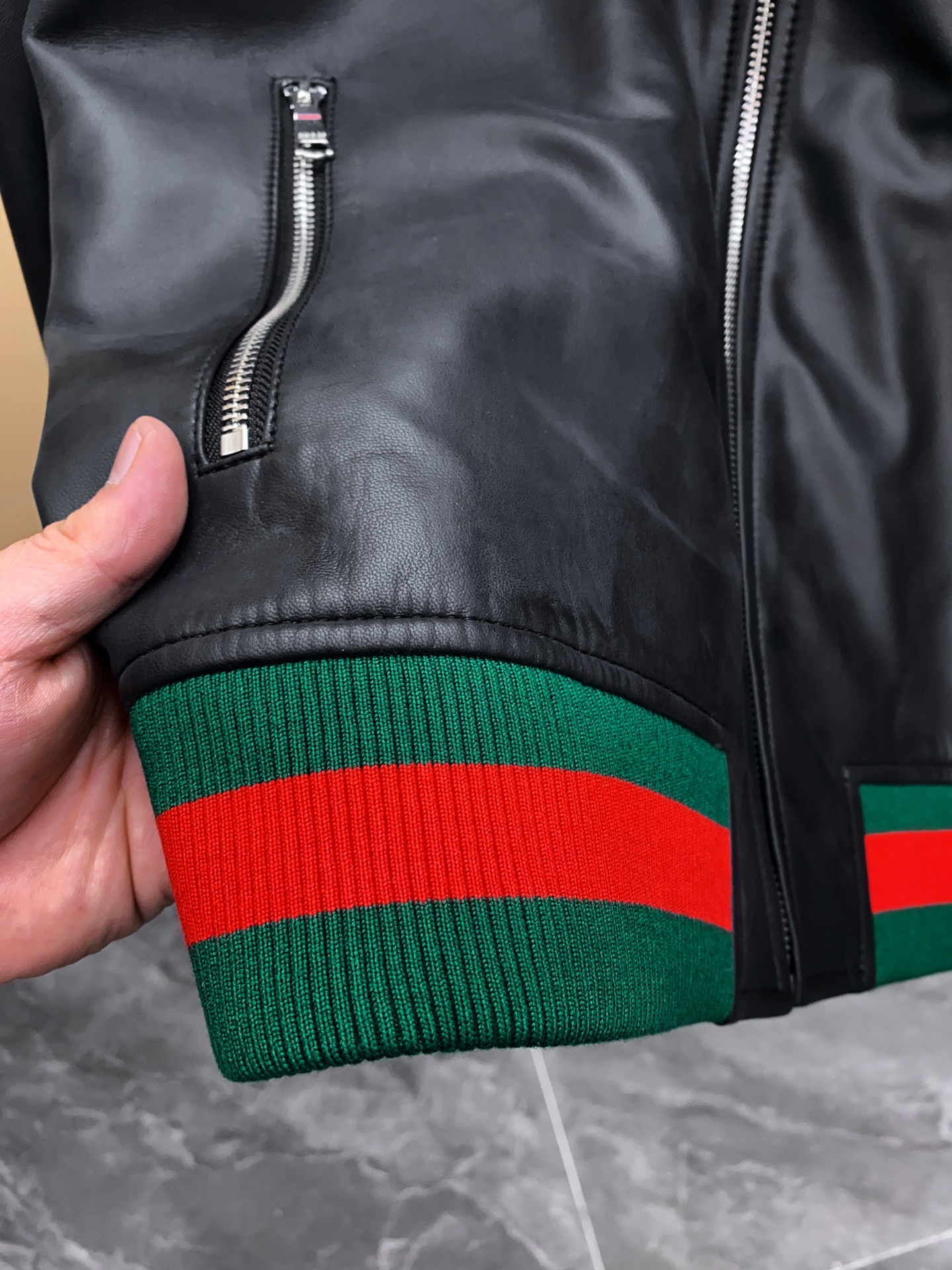 P1260 Gucci古驰纳帕皮飞行员夹克外套 。柔软纳帕皮革 + 羊毛棉混纺针织,质感与舒适度
外层选用柔软细腻的黑色纳帕羊皮,皮质纹理自然细腻,触感顺滑,兼具挺括感与柔韧度。表面光泽均匀,展现低调奢华的高级质感。 
针织拼接工艺 袖子与背部采用羊毛与棉混纺针织面料,针脚密实且富有弹性。在保持舒适穿着体验的同时,增强衣身的透气性与灵活性。立体剪裁版型直筒修身飞行员夹克版型,肩线挺括,身形利落。拼接工艺自然过渡 兼顾运动风与都市感。
门襟配备金属拉链,顺滑耐用。 袖口与下摆采用罗纹针织,并点缀 经典红绿织带,彰显标志性元素。 内里采用光滑里布拼接针织,穿着顺滑舒适。增添保暖性与耐穿性。