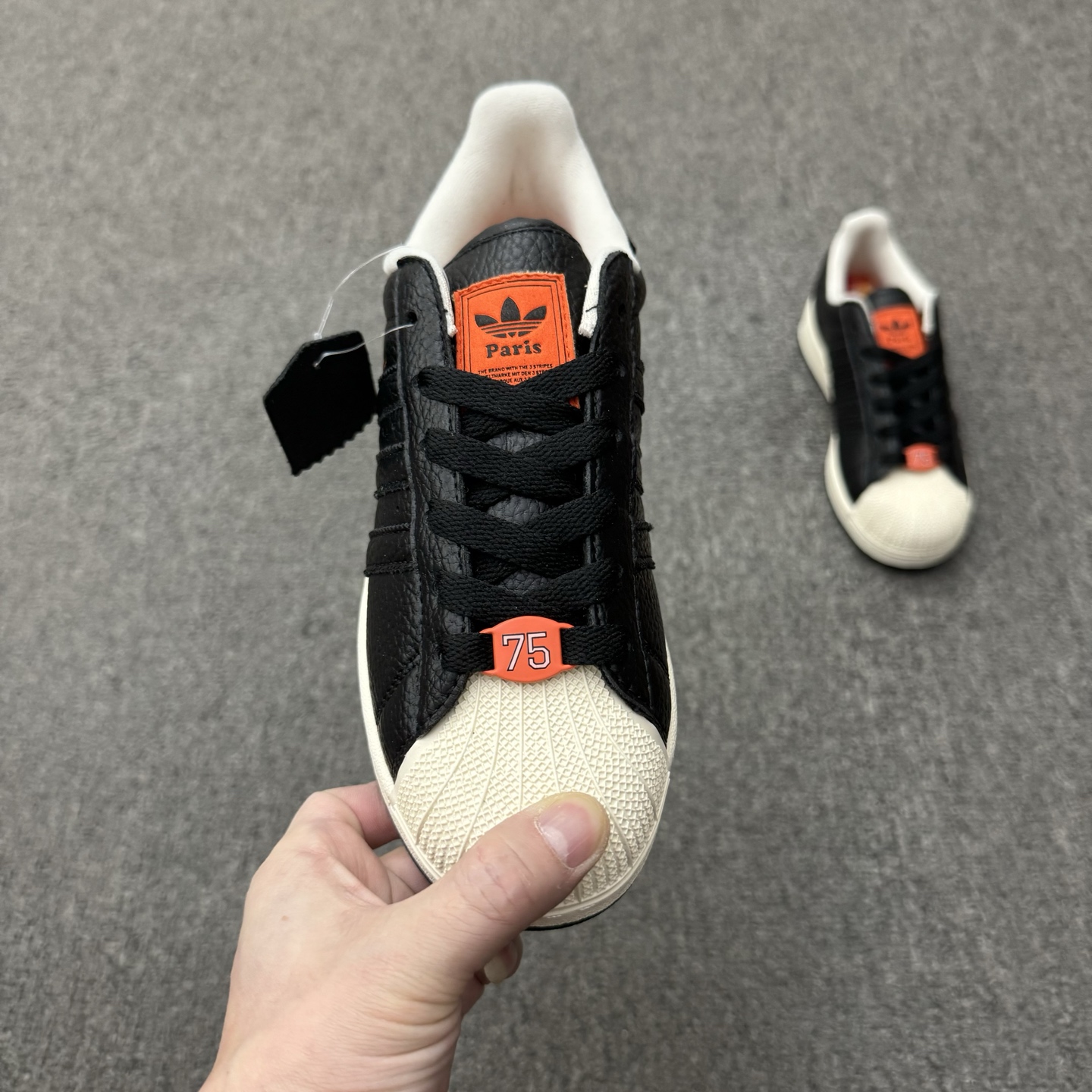 【💰💰110】（35.5-45半）
Adidas Original Superstar 2 "Paris" 阿迪达斯 低帮 巴黎联名 黑橙 男女同款 贝壳头2代防滑耐磨轻便低邦板鞋 鞋身整体以黑色皮革打造，搭配经典白色贝壳头，鞋身两侧经典锯齿三条杠Logo采用黑色点缀，侧身Superstar字样橙色压印妆点，鞋舌橙色底色上以三叶草Logo及Paris字样呈现，瞬间提升鞋子的时尚程度，后跟装饰片辅以黑色皮革制作并带有白色三叶草Logo及Paris字样，外加法国75数字的鞋带饰品致敬巴黎区号，最后以白色中底搭配生胶色橡胶外底完善整体设计收尾。
货号：JQ3223
尺码：35¹/₃ 36 36²/₃ 37¹/₃ 38 38²/₃ 39¹/₃ 40 40²/₃ 41¹/₃ 42 42²/₃ 43¹/₃ 44 44²/₃ 45¹/₃