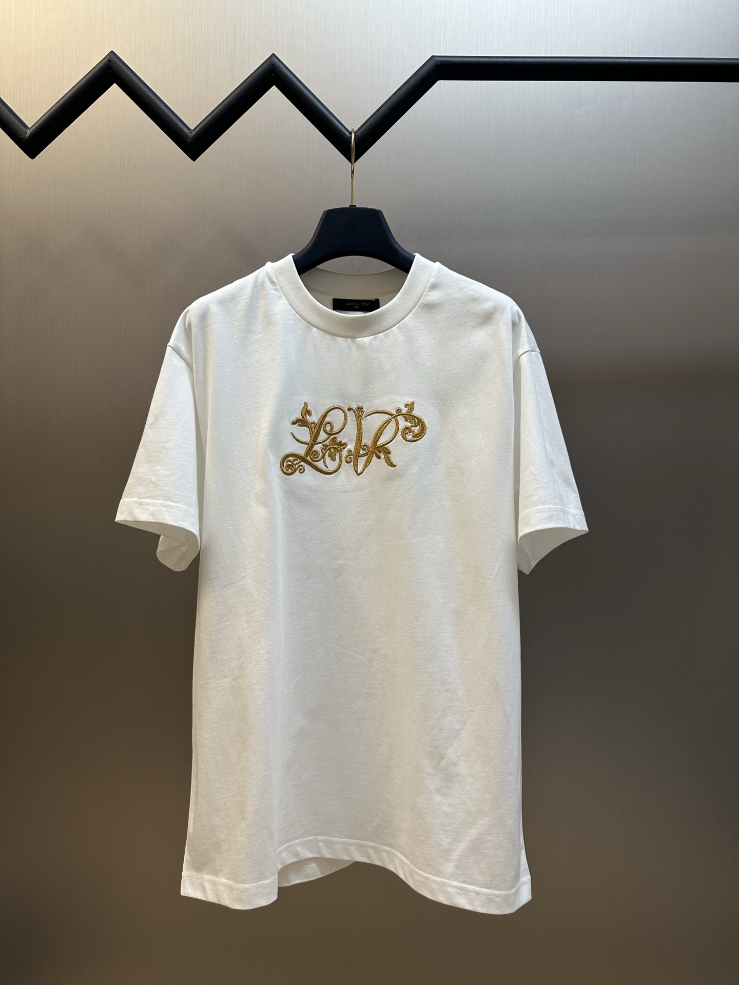 NO:845759,LU gold flower embroidered round neck short sleeves Soft cotton knitted fabric, made of 15-count special-spun compact cotton, with a weight of 230g/㎡; with 32-count 1×1 rib; a weight of 340g/㎡. The fabric is a medium-thick knitted fabric, with a thicker 15-count yarn, a strong fabric texture, and a crisp texture. The cotton fiber cross-section of the fabric is round, which significantly enhances the gloss and smoothness. Light energy improves the adsorption uniformity of the dye, making the fabric background color fuller and more uniform. It is then stretched and set by a high-temperature setting machine, and the warp and weft yarns are restored to straightness through tension control, eliminating wrinkles and internal stress. The 32-count rib collar retracts to its optimal elastic state, and the tight structure of the rib is fully stabilized during this process, forming a collar that fits well without being stretched. The gold thread flower embroidery is shown in the center. The gold thread needle embroidery technique is a fine and decorative embroidery technique. It is made of metal alloy threads with tight stitches, bright colors and high gloss. The gold thread is wound on the spool to ensure that the thread ends are neat and easy to control, forming a tight stitch. The embroidery is very fine and the stitches are dense, the fonts are clear and the lines and three-dimensional sense are strong. The subtle visual texture of the gold thread makes the embroidery appear full and three-dimensional. The overall stitching is fine and even, and the fineness and texture are consistent. The golden embroidery forms a material contrast with the cotton base fabric, which not only highlights the tone of the floral pattern, but also maintains the restoration and refinement of the overall design. It adopts a box-shaped straight cut, with the shoulder line accurately falling on the acromion point or slightly moving outward. The body is H-shaped straight, with similar bust, waist and hem circumferences, and no waist design. The box-shaped cut is characterized by square outlines and neat lines. The drape and support of the fabric ensure that the box-shaped structure will not collapse due to wearing. The overall structure and comfort are taken into account under the loose frame, making the central golden flower embroidery the visual center of gravity, forming a balance with the neat box-shaped silhouette. sizes m l,,T-shirt,19860909LU 金花刺绣圆领短袖 柔软棉质针织面料,采用15支特纺紧密棉,克重为230g/㎡；搭配32支1×1罗纹；克重为340g/㎡.面料属于中厚型针织面料,15支纱线较粗,织物肌理感强,质地挺括.面料棉纤维截面圆润,显著增强光泽度与平滑感,丝光能提升染料的吸附均匀性,使面料底色更加饱满匀净.后经高温定型机拉幅定形,通过张力控制使经纬纱恢复平直,消除褶皱与内应力.32支罗纹的领口回缩至最佳弹性状态,罗纹的紧实结构在此过程中充分稳定,形成贴合而不勒的领型. 中央呈现金丝花卉刺绣,金线挨针刺绣工艺是一种精细且富有装饰性的刺绣技法,使用金属合金线制成,线迹紧密、色彩鲜艳、光泽度高.将金线绕在线轴上,确保线头整齐且易于操控,形成紧密的线迹.刺绣非常精细针法绵密,字体清晰且线条感和立体感强. 金线的微妙视觉质感使绣体呈现饱满立体的效果,整体针脚细密均匀,精细度与质感保持一致.金色刺绣与棉质底布形成材质对比,既突出了花卉图案的色调,又保持了整体设计的还原与精致. 采用箱型直身剪裁,肩线精准落在肩峰点或略微外移,衣身呈H型直筒,胸围、腰围、下摆围度相近,无收腰设计.箱型剪裁的特点是轮廓方正、线条利落,面料的垂坠性与支撑力保证了箱型结构不会因穿着而塌陷.整体在宽松框架下兼顾了结构感与舒适度,使中央金花刺绣成为视觉重心,与利落的箱型廓形形成平衡 sizes m l,,T-shirt,,Men's clothing
