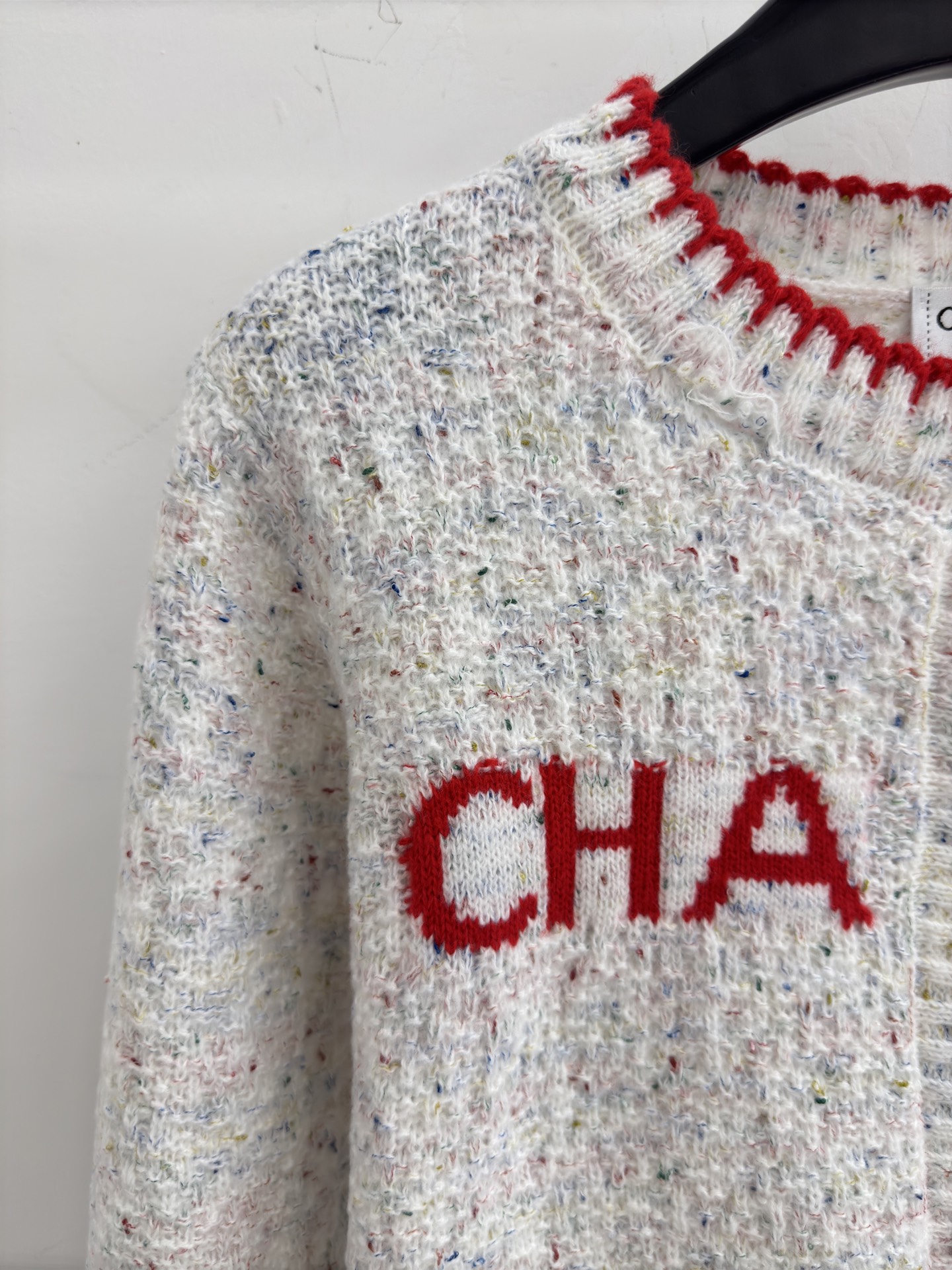 Chanel 字母针织开衫‌，灰白色混纺针织面料，带有细微的彩色杂点纹理，质感柔软且透气，胸前红色字母提花字体简约