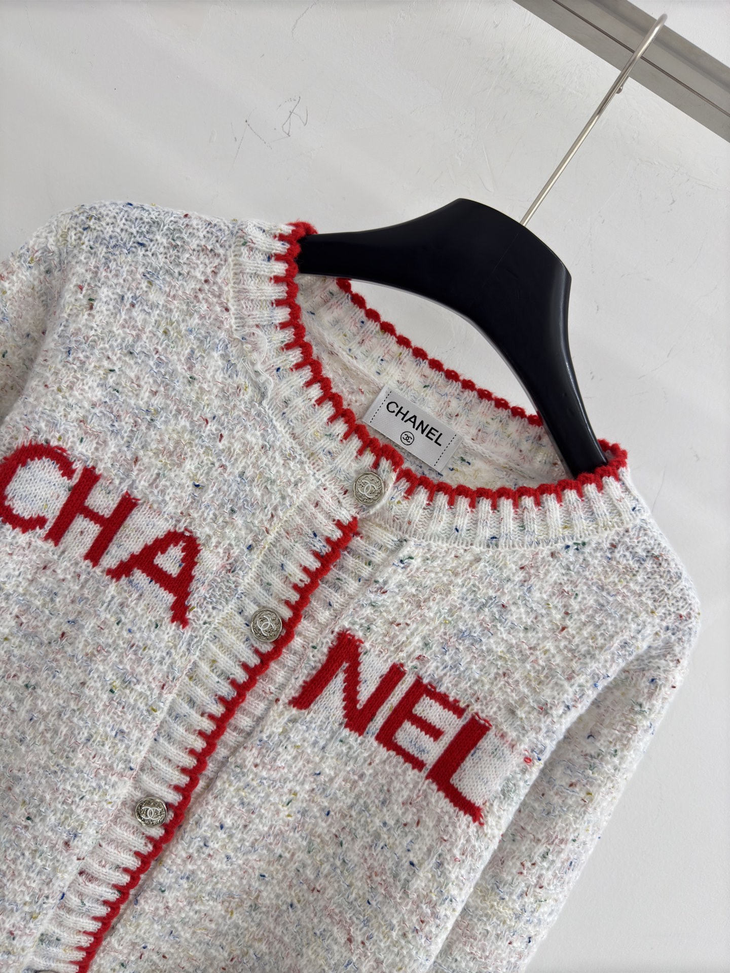Chanel 字母针织开衫‌，灰白色混纺针织面料，带有细微的彩色杂点纹理，质感柔软且透气，胸前红色字母提花字体简约