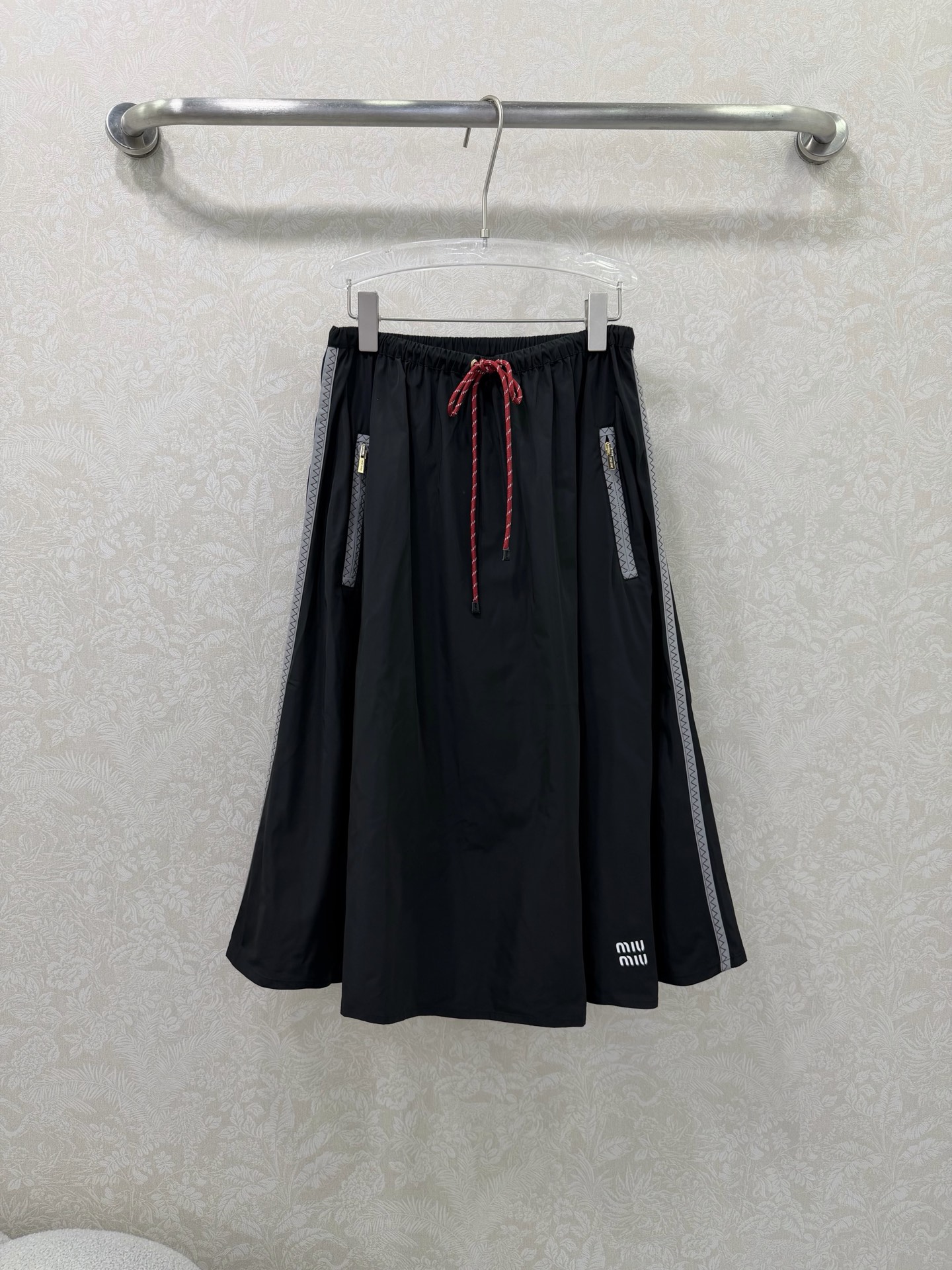 NO:744622,miumi*26 new side webbing large swing umbrella skirt is an iconic item of miu style!  The skirt is spliced with gray webbing on both sides, with delicate zigzag patterns on the edges, which visually elongates the legs vertically and enhances the sense of sports and leisure. The front pockets are equipped with customized gold zippers, which enhance the light luxury texture, are both practical and decorative, and are convenient for placing small items. The elastic waist + contrasting drawstring design (blue and white drawstrings for the navy model, red and white drawstrings for the black model) are easy to put on and take off without picking up the waistline, adding a sense of liveliness.  The high-waisted A-line version with a wide hem and natural draping can effectively hide the fat on the crotch and thighs. It is friendly to all body types. The skirt is flexible and elegant when walking. The skirt is printed with the white 'miu mi*' letter logo, which low-key highlights the style attribute and makes it highly recognizable. 2 colors and 3 codes SMLR, Miu Miu-clothing, miumiu, skirts,19860909miumi*26新款侧织带大摆伞裙 miu系穿搭的标志性单品！裙身两侧拼接灰色织带,边缘带有精致锯齿纹,视觉上纵向拉长腿型,同时强化运动休闲感.前侧口袋配有定制金色拉链,提升轻奢质感,兼具实用性与装饰性,方便放置小物件.松紧腰头+撞色抽绳设计（藏蓝款为蓝白抽绳,黑色款为红白抽绳）,穿脱方便且不挑腰围,增加活泼感 高腰A字大摆版型,自然垂坠散开,有效藏住胯部与大腿赘肉,对各种身材都很友好,走路时裙摆灵动飘逸 裙身印有白色“miu mi*”字母标识,低调点明风格属性,辨识度拉满. 2色3码SMLR,缪缪-服装,miumiu,skirts,,Women's clothing