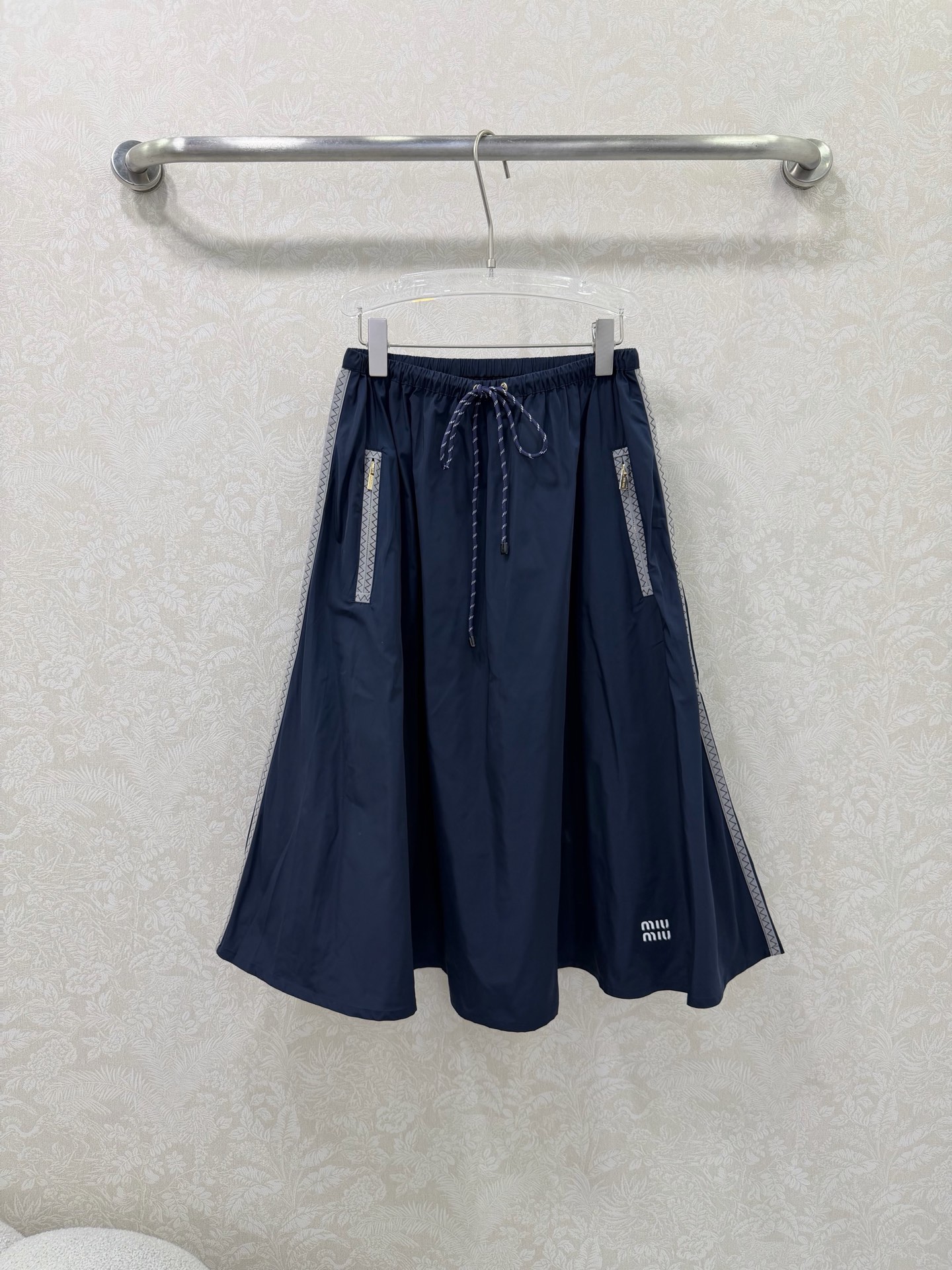 NO:744638,miumi*26 new side webbing large swing umbrella skirt is an iconic item of miu style!  The skirt is spliced with gray webbing on both sides, with delicate zigzag patterns on the edges, which visually elongates the legs vertically and enhances the sense of sports and leisure. The front pockets are equipped with customized gold zippers, which enhance the light luxury texture, are both practical and decorative, and are convenient for placing small items. The elastic waist + contrasting drawstring design (blue and white drawstrings for the navy model, red and white drawstrings for the black model) are easy to put on and take off without picking up the waistline, adding a sense of liveliness.  The high-waisted A-line version with a wide hem and natural draping can effectively hide the fat on the crotch and thighs. It is friendly to all body types. The skirt is flexible and elegant when walking. The skirt is printed with the white 'miu mi*' letter logo, which low-key highlights the style attribute and makes it highly recognizable. 2 colors and 3 codes SMLR, Miu Miu-clothing, miumiu, skirts,19860909miumi*26新款侧织带大摆伞裙 miu系穿搭的标志性单品！裙身两侧拼接灰色织带,边缘带有精致锯齿纹,视觉上纵向拉长腿型,同时强化运动休闲感.前侧口袋配有定制金色拉链,提升轻奢质感,兼具实用性与装饰性,方便放置小物件.松紧腰头+撞色抽绳设计（藏蓝款为蓝白抽绳,黑色款为红白抽绳）,穿脱方便且不挑腰围,增加活泼感 高腰A字大摆版型,自然垂坠散开,有效藏住胯部与大腿赘肉,对各种身材都很友好,走路时裙摆灵动飘逸 裙身印有白色“miu mi*”字母标识,低调点明风格属性,辨识度拉满. 2色3码SMLR,缪缪-服装,miumiu,skirts,,Women's clothing