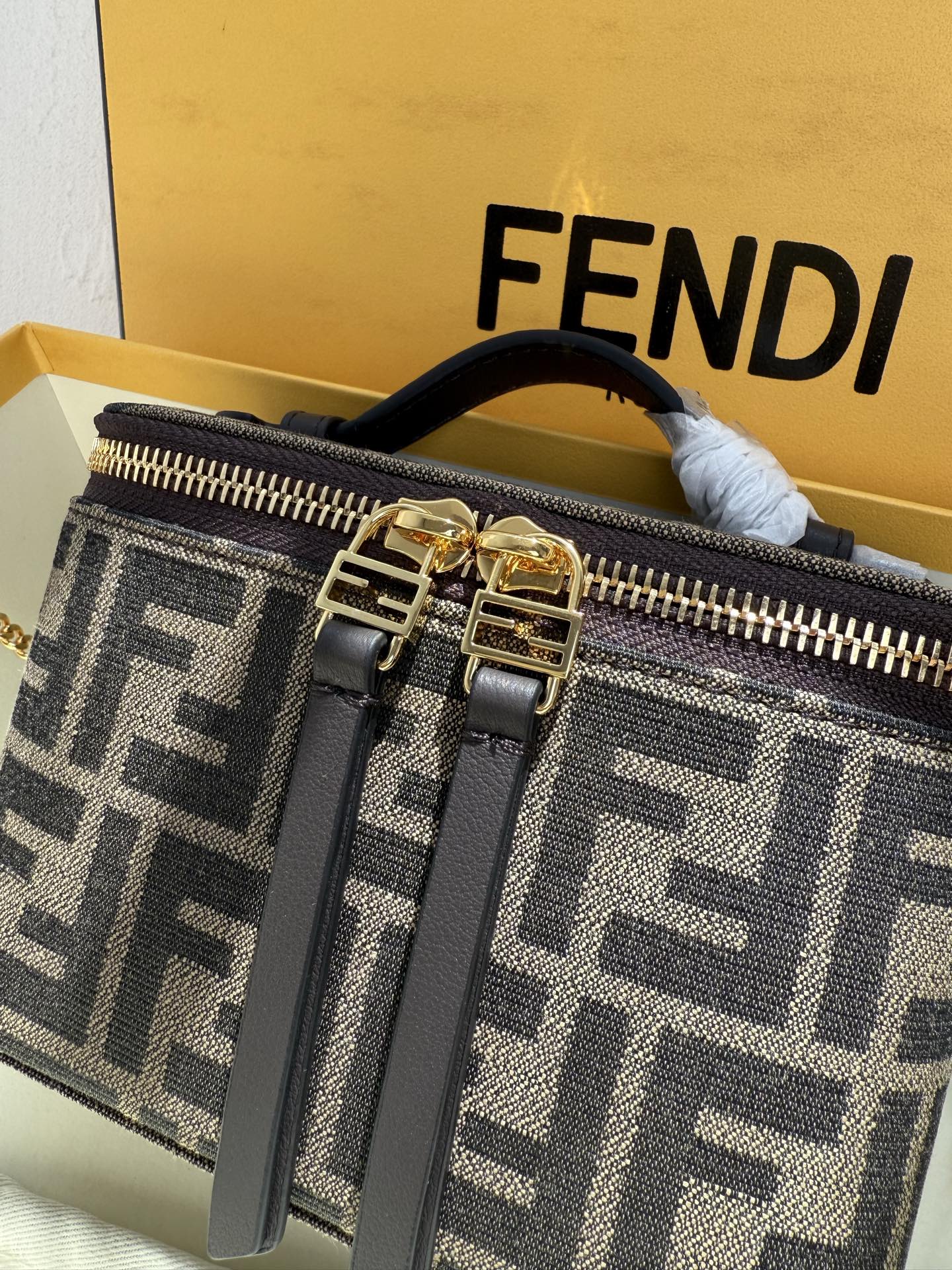 Fendi 廓形挺立的小巧型Vanity手袋采用双滑动拉链开合设计，饰有FF细节和皮革拉链头。烟草棕FF提花布料材质