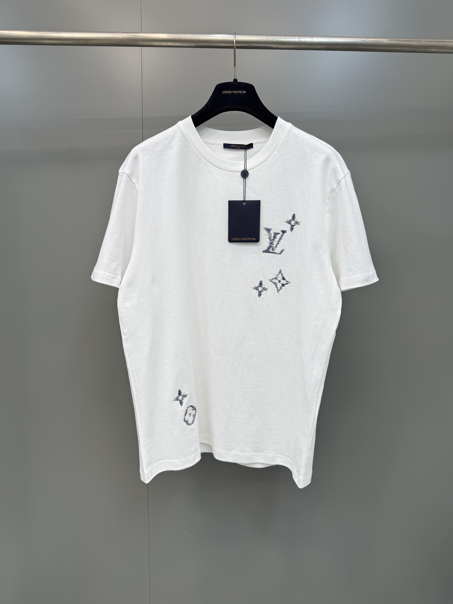 NO:821124,Lujia 26ss new embroidered round neck short-sleeved T-shirt is made of 12-count heavyweight combed pure cotton plain fabric, weighing 230g. The fabric texture is full and thick, with strong structural support and skin-friendly breathability. It has been pre-shrunk and enzymatically pre-washed. It has excellent dimensional stability and can effectively resist the risk of deformation and wrinkles caused by daily washing. It can still maintain a neat silhouette after long-term wear.  The version adopts a slightly loose straight cut, and the neckline is a double-needle ribbed round collar. The overlocking process is used to enhance the collar's stretch resistance, making it less likely to loosen and deform after long-term wear. The brand's Monogram logo scattered on the front and back panels is made of precision flat embroidery technology and is embroidered in blue.  The high-twist cotton thread is embroidered through computer plate making, using a unique imitation hand embroidery random stitching technique to simulate the raw edge texture of hand-painted graffiti. The stitches are fine and neat, presenting a layered three-dimensional relief texture on the plain base fabric, breaking the regularity of traditional embroidery.  The cuffs and hem are rolled using flat-turn double-needle hemming, with clean and neat lines. It perfectly integrates the brand's classic elements with modern deconstructed aesthetics, making it both practical and fashionable for daily wear. Louis Vuitton-clothing, louis vuitton, louis vuitton, louis vuitton, T-shirt, tees, t-shirt,19860909驴家26ss新款刺绣圆领短袖T恤,采用12支重磅精梳纯棉平纹面料织造,克重230g,布面肌理饱满厚实,兼具强韧结构支撑力与亲肤透气性,经预缩定型与酵素预洗处理,尺寸稳定性优异,可有效抵御日常洗涤带来的变形、起皱风险,久穿仍保持利落廓形 版型上采用微宽松直身剪裁,领口为双针罗纹圆领,配合锁边工艺强化领型抗拉伸性,久穿不易松垮变形；前后幅散落的品牌Monogram标识采用精密平绣工艺,以蓝色高捻棉线通过电脑制版绣制,采用独特的仿手绣乱针走线技法,模拟手绘涂鸦的毛边肌理,针脚细密工整,在素色底布上呈现出层次丰富的立体浮雕质感,打破传统刺绣的规整感 袖口与下摆采用平车双针卷边处理,线条干净利落,将品牌经典元素与现代解构美学完美融合,兼具日常实穿性与潮流辨识度,路易威登-服装,louis vuitton,louis vuitton,louis vuitton,T-shirt,tees，t-shirt,,Women's clothing