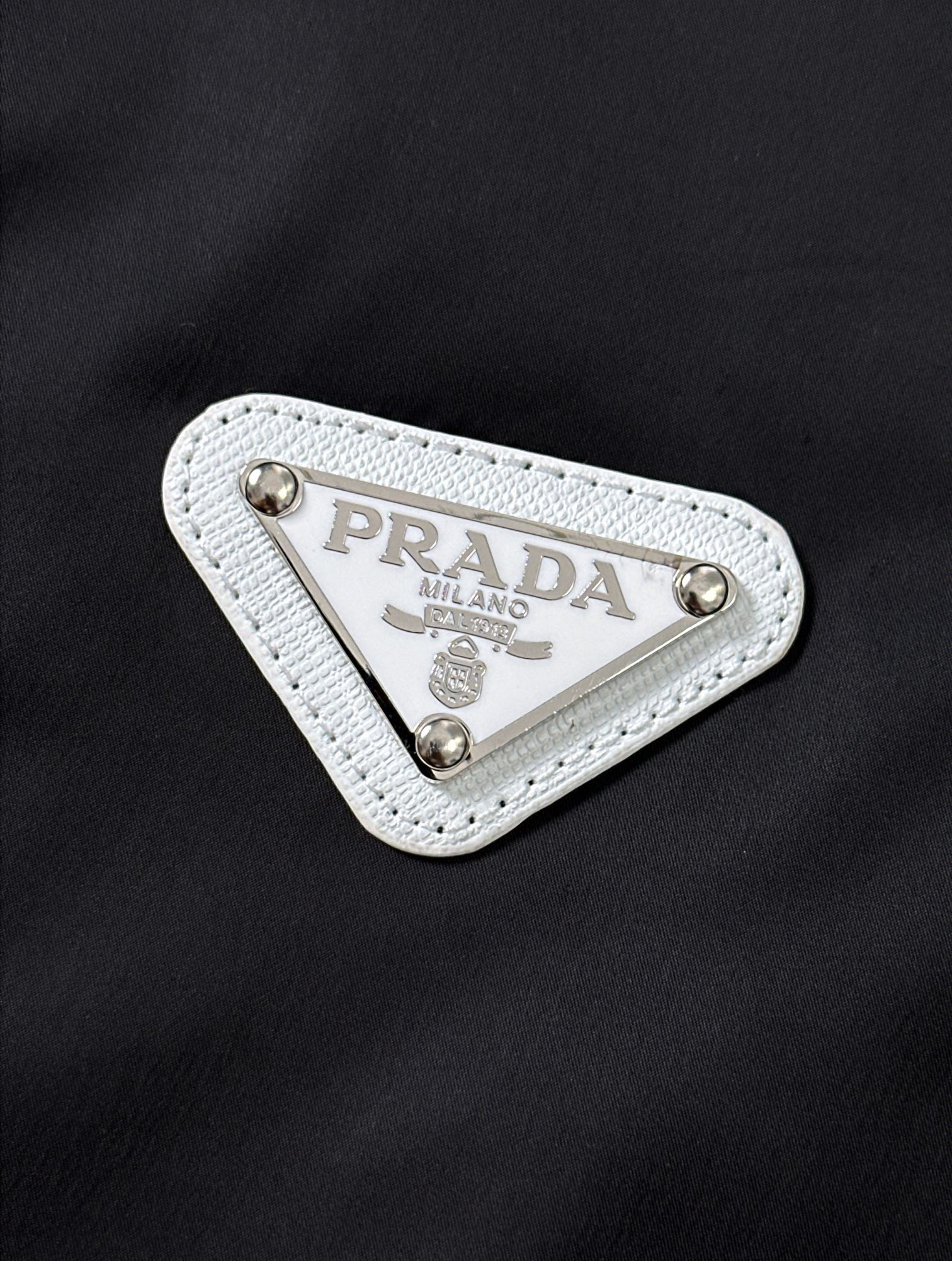 P320  896 PRA……普拉达 24新品连帽夹克！先上先卖，抢占先机。PRADA/普拉达 新品连帽风衣外套。这款轻质尼龙夹克和正面拉链，下摆特殊设计，颇具休闲装设计特色。胸前拼接白色门襟。演绎品牌风范。连帽，正面拉链式口袋，饰有涂珐琅三角形金属徽标，正面拉链拉合，聚酰胺纤维面料。黑色 尺码：48-56