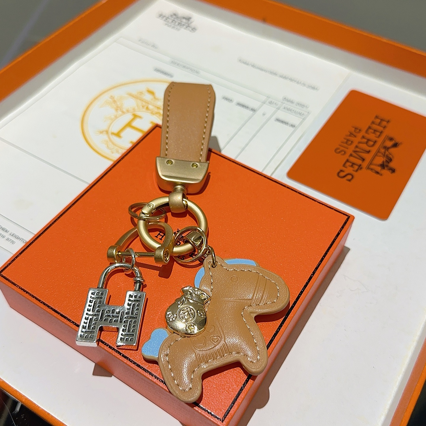 Chaveiro com pingente de cavalo Hermes, couro bronzeado com detalhes em azul claro, hardware dourado fosco, tom prateado, pingente H, comprimento total de 10 cm