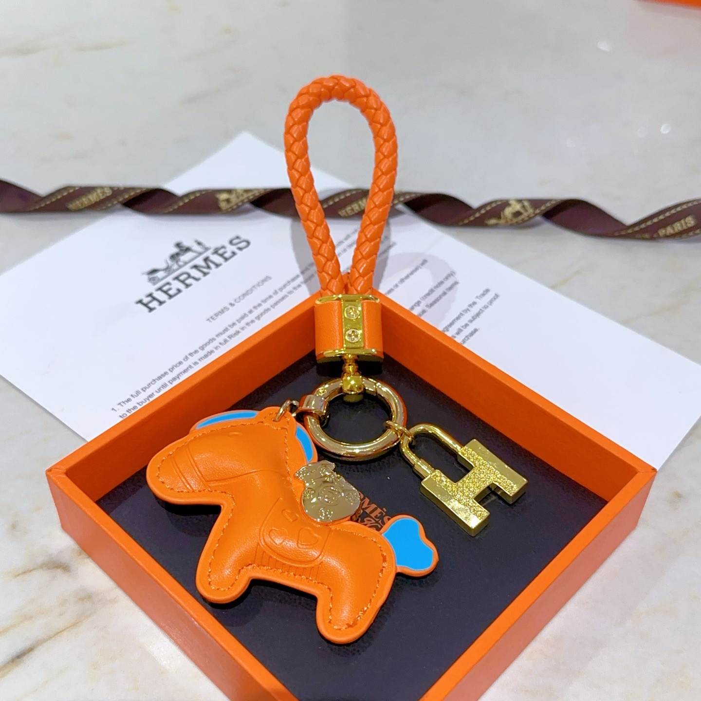 Ciondolo Hermes Rodeo H Lock Key Charm Ciondolo cavallo in pelle arancione intrecciata in pelle intrecciata arancione in metallo color oro con accenti blu, lunghezza totale 10 cm
