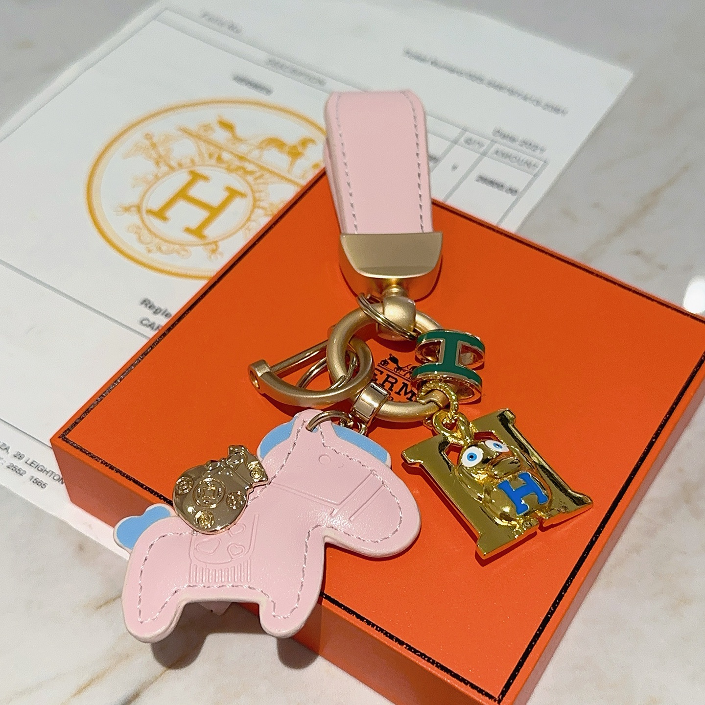 Hermes Rodeo H Logo Cartoon Charm Chaveiro pingente Couro rosa com detalhes em azul claro metal dourado detalhes em esmalte verde e azul 10 cm de comprimento total