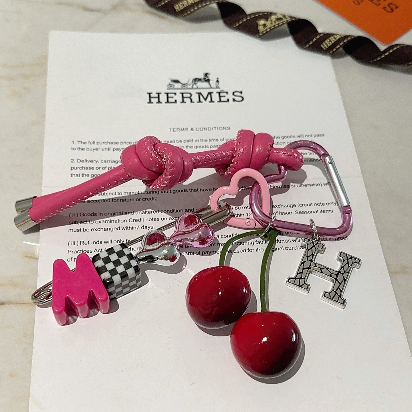 Hermes Cherry H Logo Knot Bag Charm Llavero colgante Cuero rosa intenso Metal en tono plateado Detalles en cereza roja Detalle a cuadros en blanco y negro Herrajes en forma de corazón rosa Tallos de cereza verde 10 cm