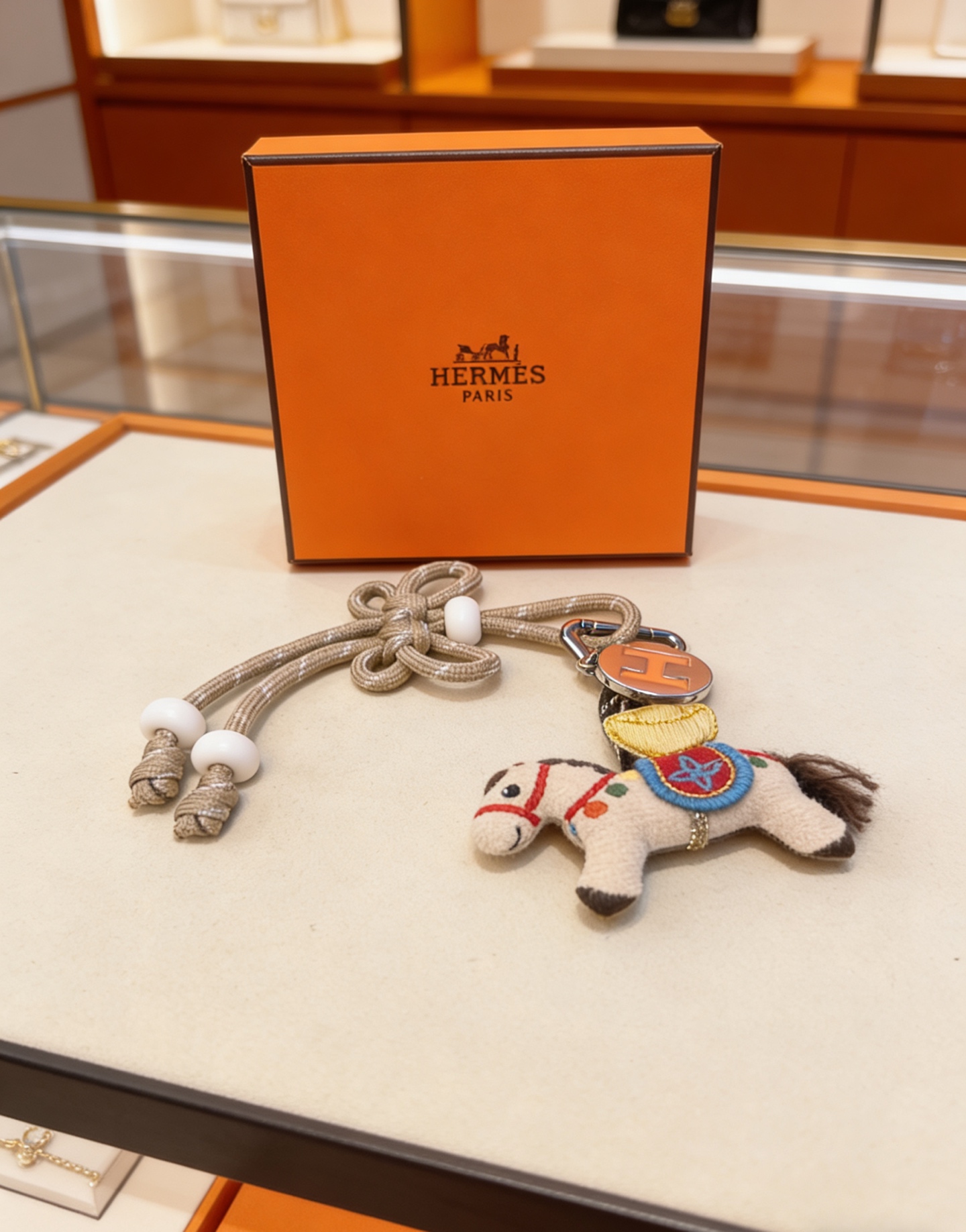 Pendentif porte-clés Hermes Rodeo Horse Charm Métal argenté avec émail orange Logo H Cordon tressé beige avec perles de résine blanches Breloque cheval en feutre blanc cassé avec accents brodés multicolores et queue de crinière marron Longueur totale 12 cm