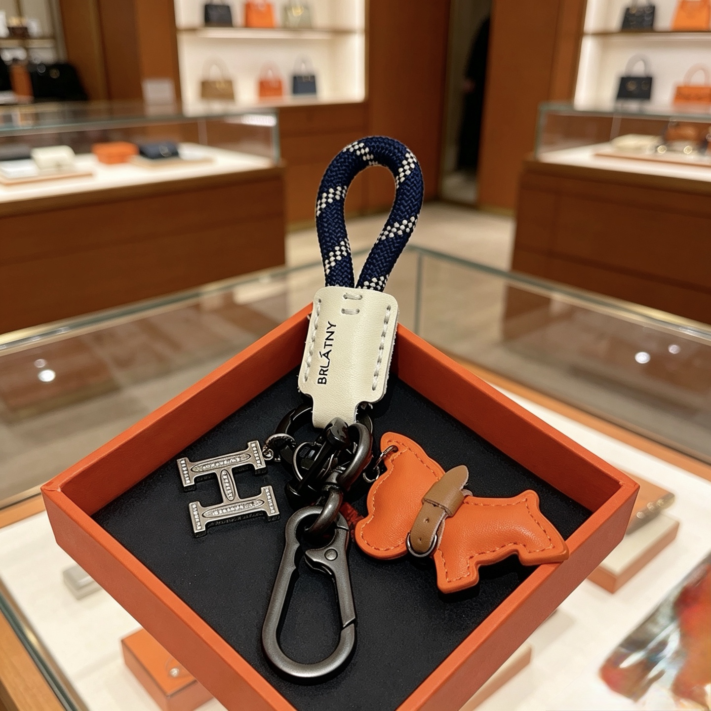 Hermes BRLATNY Puppy H Charm Llavero colgante Herrajes de bronce azul marino con correa tejida a cuadros blancos Etiqueta de cuero blanquecino Charm de cachorro de cuero naranja con collar de cuero marrón Detalles de cristal transparente en el dije H 10 cm de longitud total