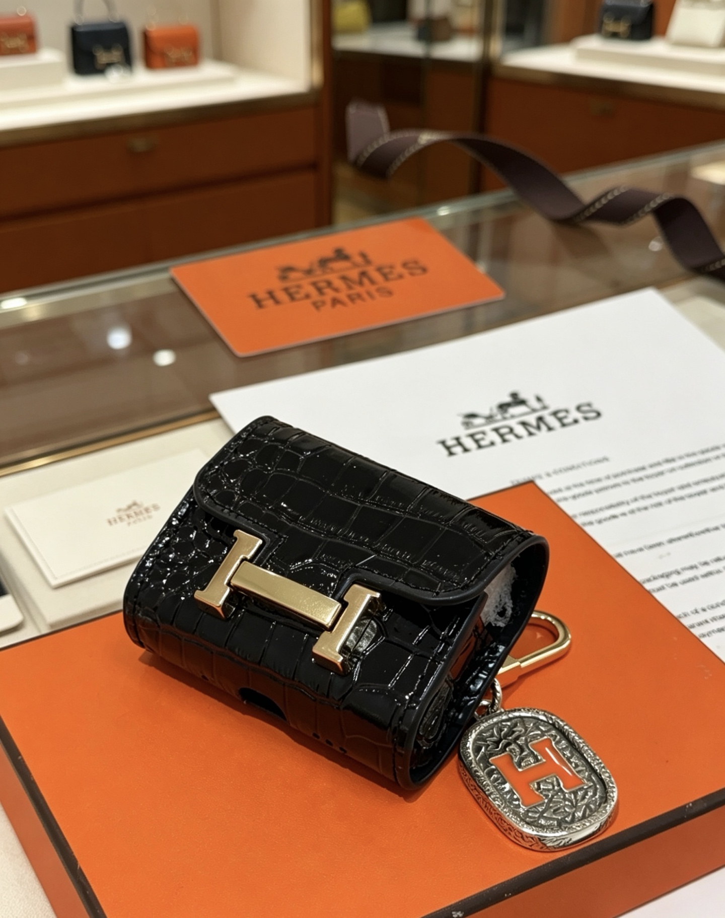 Hermes Constance AirPods Case Bag Charm pendentif Cuir verni noir embossé crocodile Quincaillerie en métal doré Charme en métal argenté avec émail orange Détail H Taille unique pour le charme AirPods standard longueur totale environ 8 cm