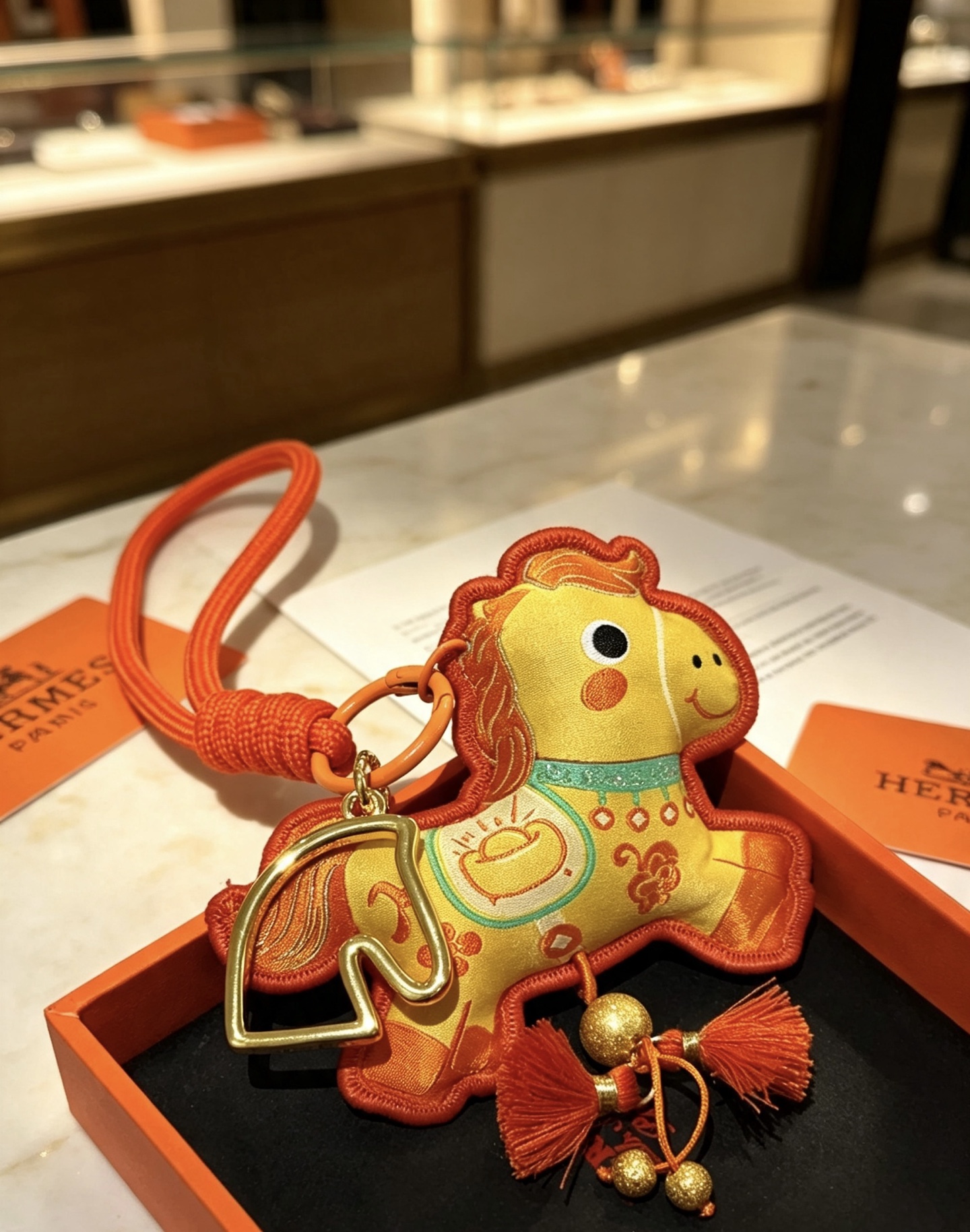 Hermes Año del caballo Colgante Lucky Horse Charm Metal en tono dorado Tela amarilla Tela y ribete naranja Detalles bordados en turquesa Borlas naranjas Longitud de la correa para la muñeca 18 cm