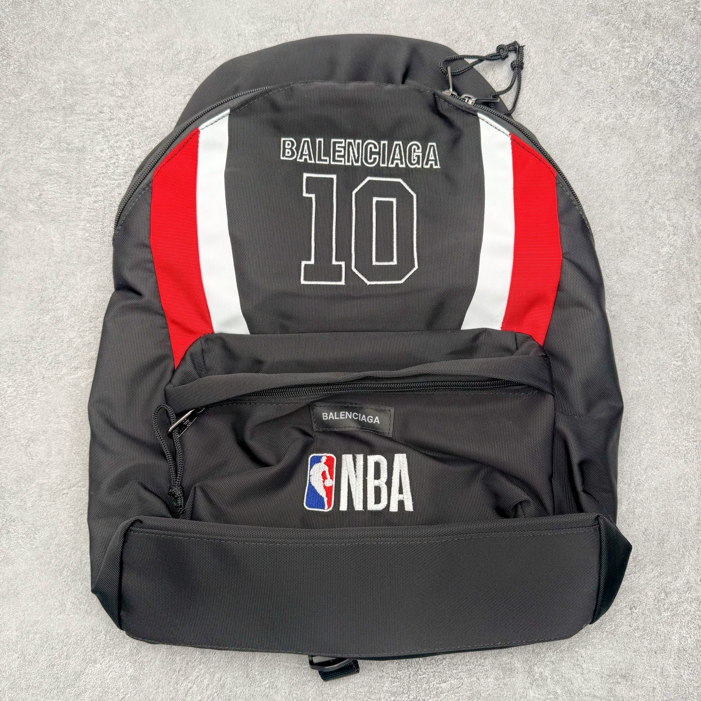 Balenciaga巴黎世家 26年最新款 NBA联名系列 刺绣标识 双肩包 当高奢时尚与篮球文化碰撞，这款双肩包便成了街头与秀场的通行证。 • 设计：以经典黑色尼龙为基底，正面醒目的“BALENCIAGA 10”字样，复刻了球衣号码的热血基因；两侧红白撞色条纹，既是对赛场元素的致敬，也是视觉焦点。 • 细节质感：前置口袋上的NBA官方徽标与BALENCIAGA织标双重认证，从材质到走线，都延续了品牌对工艺的极致追求。 • 风格适配：无论是日常出街、球场边的随性搭配，还是作为潮流收藏，它都能轻松hold住多元场景，让运动精神与高级时装感无缝衔接 材质：高档锦纶帆布 尺寸：48cm*31cm*20cm-选品中心