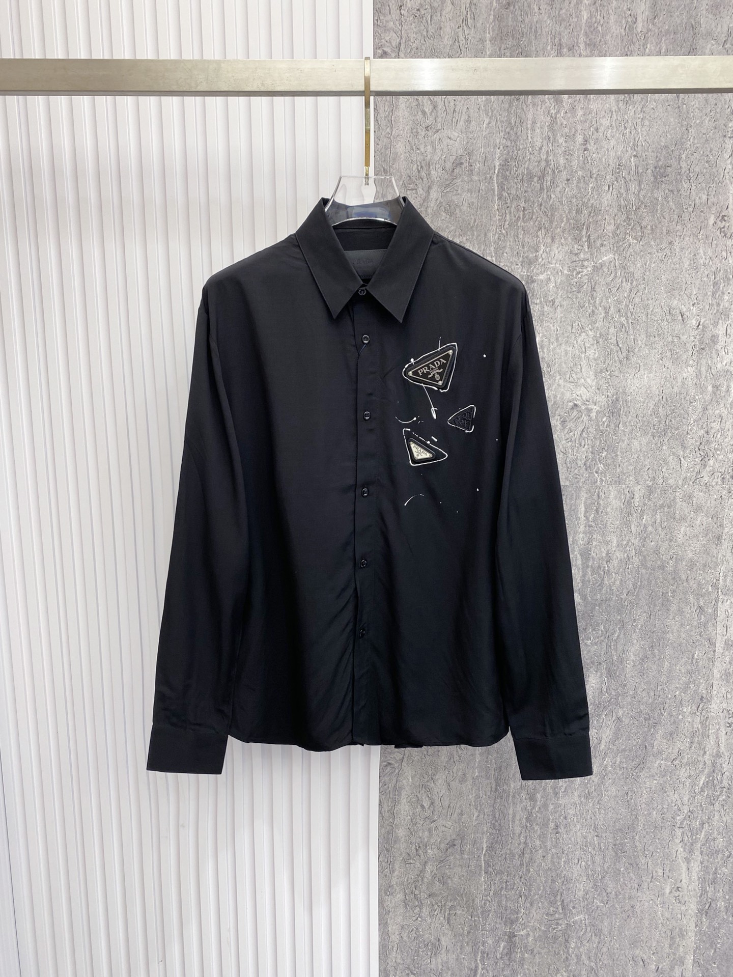 NO:842781,Prada Prada  2026ss spring and summer graffiti triangle logo black shirt, using street graffiti elements and minimalist tailoring to interpret the dark high-end relaxed aesthetics. Made of high-draping matte satin fabric, it feels silky and skin-friendly, has a low-key soft light texture, and the upper body is draped with  shape; the body adopts a neat straight silhouette, and the graffiti-style triangle logo on the chest breaks the dullness and injects street vitality. Whether worn alone to create a cool commuting style, or layered with a suit to create a business elite feel, it can easily create an effortless high-quality outfit.  Size 48-56,,prada,prada,shirts,business suit,19860909Prada 普拉达 2026ss春夏涂鸦三角标黑衬衫,以街头涂鸦元素与极简剪裁诠释暗黑系高阶松弛美学.甄选高垂坠哑光缎面面料打造,触感丝滑亲肤,自带低调柔光质感,上身垂坠有型；衣身采用利落直筒版型,胸口涂鸦风三角标打破沉闷,注入街头活力.无论是单穿打造酷感通勤风,还是叠穿西装演绎商务精英感,都能轻松塑造不费力的高级质感穿搭. 码数48-56,,prada,prada,shirts,business suit,,Men's clothing