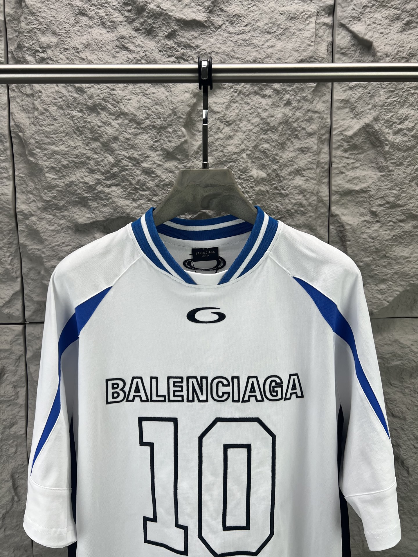 Balenciaga BALENCI*GA × NBA SS26联名T恤 采用科技网眼面料 经透气排汗工艺处理 兼具运动功能性与 廓形 衣身主体为黑色 肩部与袖口拼接撞色饰带 采用精密车缝工艺 线条利落 正面“BALENCIAGA”字样与数字“10”采用立体植绒或贴布绣工艺 边缘锁边牢固 不易脱线 领口为罗纹圆领 采用高弹氨纶混纺材质 久穿不易变形 下摆与袖口采用双针锁边工艺 提升整体耐用性 衣身印有NBA 标识 采用环保胶印工艺 图案清晰牢固 整体设计融合高端时装与运动元素 兼顾舒适穿着与视觉张力 适合潮流穿搭与收藏 整体走线平直细腻 面料舒适柔软 颜色一致无色差 能与同款搭配营造 造型 驾驭各种场合 定制辅料 三标齐全