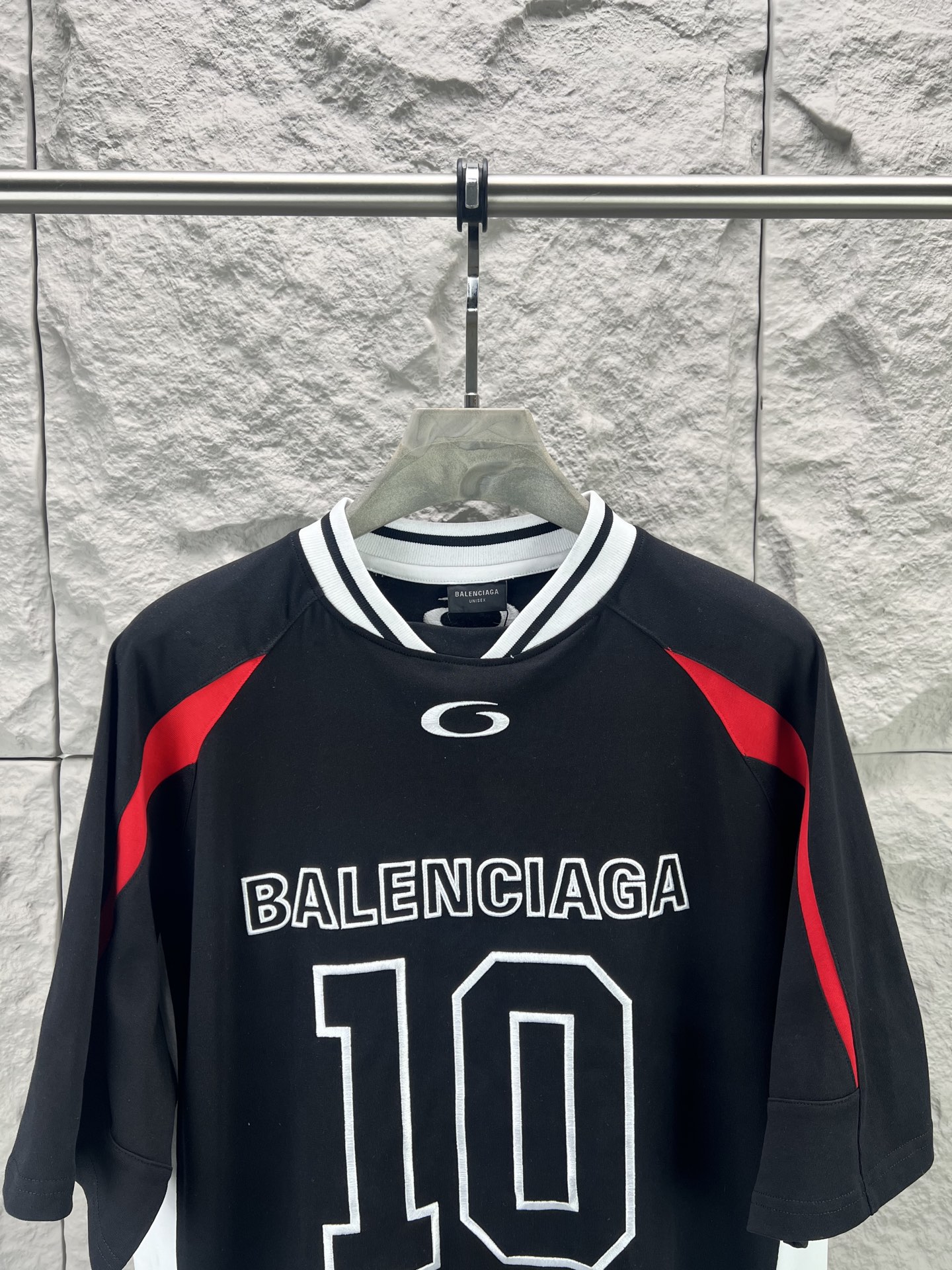 Balenciaga BALENCI*GA × NBA SS26联名T恤 采用科技网眼面料 经透气排汗工艺处理 兼具运动功能性与 廓形 衣身主体为黑色 肩部与袖口拼接撞色饰带 采用精密车缝工艺 线条利落 正面“BALENCIAGA”字样与数字“10”采用立体植绒或贴布绣工艺 边缘锁边牢固 不易脱线 领口为罗纹圆领 采用高弹氨纶混纺材质 久穿不易变形 下摆与袖口采用双针锁边工艺 提升整体耐用性 衣身印有NBA 标识 采用环保胶印工艺 图案清晰牢固 整体设计融合高端时装与运动元素 兼顾舒适穿着与视觉张力 适合潮流穿搭与收藏 整体走线平直细腻 面料舒适柔软 颜色一致无色差 能与同款搭配营造 造型 驾驭各种场合 定制辅料 三标齐全