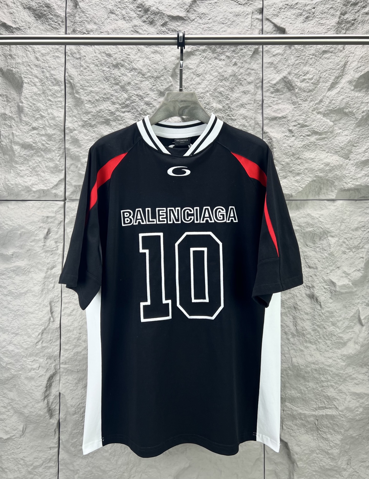 Balenciaga BALENCI*GA × NBA SS26联名T恤 采用科技网眼面料 经透气排汗工艺处理 兼具运动功能性与 廓形 衣身主体为黑色 肩部与袖口拼接撞色饰带 采用精密车缝工艺 线条利落 正面“BALENCIAGA”字样与数字“10”采用立体植绒或贴布绣工艺 边缘锁边牢固 不易脱线 领口为罗纹圆领 采用高弹氨纶混纺材质 久穿不易变形 下摆与袖口采用双针锁边工艺 提升整体耐用性 衣身印有NBA 标识 采用环保胶印工艺 图案清晰牢固 整体设计融合高端时装与运动元素 兼顾舒适穿着与视觉张力 适合潮流穿搭与收藏 整体走线平直细腻 面料舒适柔软 颜色一致无色差 能与同款搭配营造 造型 驾驭各种场合 定制辅料 三标齐全