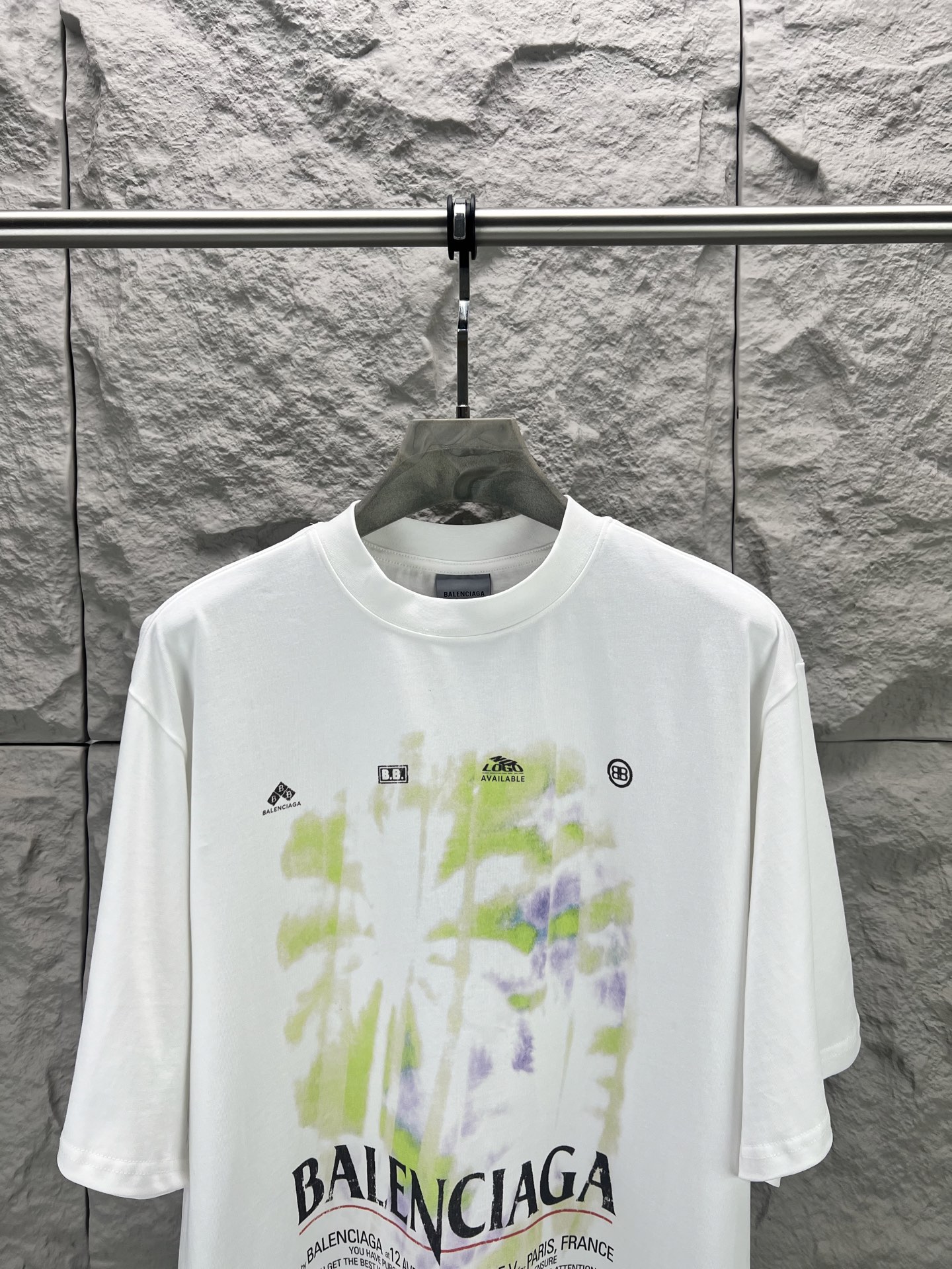Balenciaga BALENCI*GA 椰树丛林圆领短袖 采用24支特纺紧密棉平纹面料 32支1×1罗纹 面料属于兼具挺括与舒适的中厚型针织 通过紧密纺技术使纤维排列平行 纱体紧致 毛羽显著减少 表面光洁平滑 面料仅经过高温定型机拉幅定形 兼具挺括性与柔软度 垂坠感适中 穿着有型不软塌 领型采用32支罗纹 属于中等偏轻的克重 触感更加柔软轻薄 与中版型的松弛感相呼应 采用渗透印花工艺搭配网点印花制作 渗透印花是一种使染料或浆料深入渗透至纤维内部的印花方式 通过特殊的助剂与压力控制 使色彩沉入面料而非浮于表面 由于采用渗透印花工艺 图案的视觉边界柔和 无胶质感 与白色底布的融合度更高 整体走线平直细腻 面料舒适柔软 颜色一致无色差 能与同款搭配营造 造型 驾驭各种场合 定制辅料 三标齐全