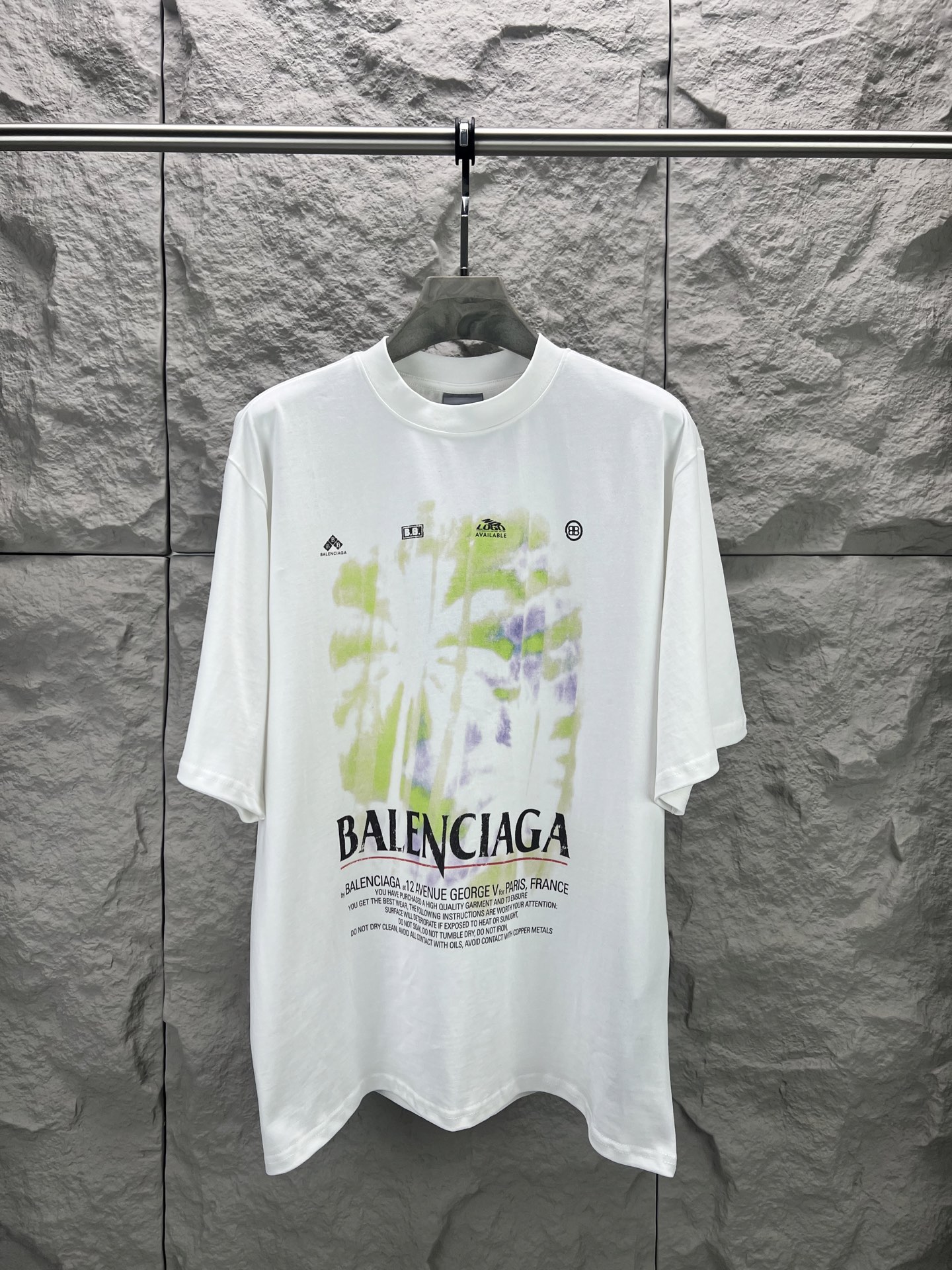 Balenciaga BALENCI*GA 椰树丛林圆领短袖 采用24支特纺紧密棉平纹面料 32支1×1罗纹 面料属于兼具挺括与舒适的中厚型针织 通过紧密纺技术使纤维排列平行 纱体紧致 毛羽显著减少 表面光洁平滑 面料仅经过高温定型机拉幅定形 兼具挺括性与柔软度 垂坠感适中 穿着有型不软塌 领型采用32支罗纹 属于中等偏轻的克重 触感更加柔软轻薄 与中版型的松弛感相呼应 采用渗透印花工艺搭配网点印花制作 渗透印花是一种使染料或浆料深入渗透至纤维内部的印花方式 通过特殊的助剂与压力控制 使色彩沉入面料而非浮于表面 由于采用渗透印花工艺 图案的视觉边界柔和 无胶质感 与白色底布的融合度更高 整体走线平直细腻 面料舒适柔软 颜色一致无色差 能与同款搭配营造 造型 驾驭各种场合 定制辅料 三标齐全