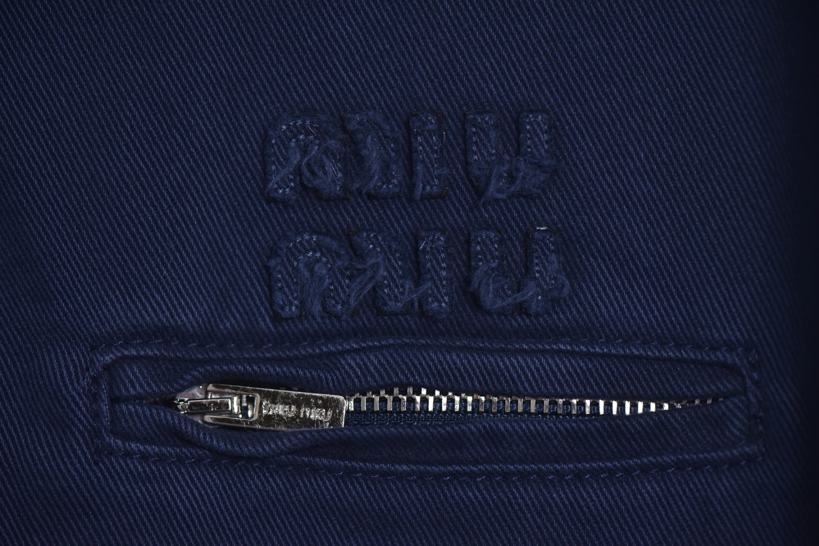 图片[3]-#Miu Miu 25Fw Miu小标法工夹棉外套 采用16OZ进口高克重牛仔面料，选用更长绒的优质棉花纺成高捻度的特规纱线，以确保最终面料的强度与最终染色的纯净度。经典的牛仔斜纹织法被精确校准，以在达成高密度的同时，保留恰到好处的柔软潜力，避免过于板硬。经纬纱的排列密度远超普通牛仔，从而获得极高的抗撕裂强度和耐磨性。定染对版色纺法工蓝，染色工程师需要反复调试靛蓝的配方，精确控制染色温度、时长和氧化次数，通过多层重复染色来模拟出时间赋予的色彩层次感。内里填充400D洗水棉，填充物为经过预洗处理的棉絮。“预洗水”工艺至关重要，它去除了棉絮的“新”感和僵硬感，使其从一开始就呈现出柔软、蓬松且略带旧化肌理的触感，确保外套穿着时立即舒适，且外观呈现出自然的蓬松感和慵懒的廓形。领部材质选用特规灯芯绒，其绒条宽度和棉绒品质均为定制，以匹配主面料的厚重质感与复古风格。胸前贴布绣花首先将设计好的图案以定制裁片的形式制作出来，再以精密缝纫的方式固定在胸前主体上。这款法工外套，是一件融合了传统工装基因与高级时装工艺的复杂作品。从独家定织定染的面料，到创新性的400D洗水棉填充结构，再到耗时费工的拼接领部与立体贴布绣花，每一个环节都设置了极高的技术和成本门槛。它不仅仅是一件外套，更是一件体现了品牌对材料学、工艺学和美学极致追求的“可穿戴艺术品”。 尺码：38 40 42-选品中心