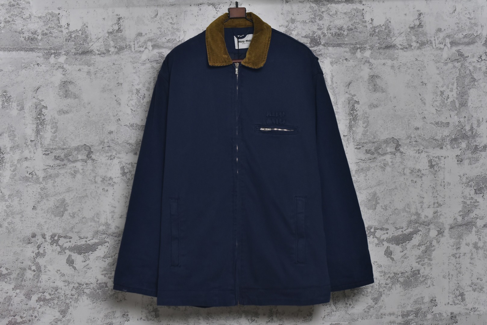 #Miu Miu 25Fw Miu小标法工夹棉外套 采用16OZ进口高克重牛仔面料，选用更长绒的优质棉花纺成高捻度的特规纱线，以确保最终面料的强度与最终染色的纯净度。经典的牛仔斜纹织法被精确校准，以在达成高密度的同时，保留恰到好处的柔软潜力，避免过于板硬。经纬纱的排列密度远超普通牛仔，从而获得极高的抗撕裂强度和耐磨性。定染对版色纺法工蓝，染色工程师需要反复调试靛蓝的配方，精确控制染色温度、时长和氧化次数，通过多层重复染色来模拟出时间赋予的色彩层次感。内里填充400D洗水棉，填充物为经过预洗处理的棉絮。“预洗水”工艺至关重要，它去除了棉絮的“新”感和僵硬感，使其从一开始就呈现出柔软、蓬松且略带旧化肌理的触感，确保外套穿着时立即舒适，且外观呈现出自然的蓬松感和慵懒的廓形。领部材质选用特规灯芯绒，其绒条宽度和棉绒品质均为定制，以匹配主面料的厚重质感与复古风格。胸前贴布绣花首先将设计好的图案以定制裁片的形式制作出来，再以精密缝纫的方式固定在胸前主体上。这款法工外套，是一件融合了传统工装基因与高级时装工艺的复杂作品。从独家定织定染的面料，到创新性的400D洗水棉填充结构，再到耗时费工的拼接领部与立体贴布绣花，每一个环节都设置了极高的技术和成本门槛。它不仅仅是一件外套，更是一件体现了品牌对材料学、工艺学和美学极致追求的“可穿戴艺术品”。 尺码：38 40 42-选品中心