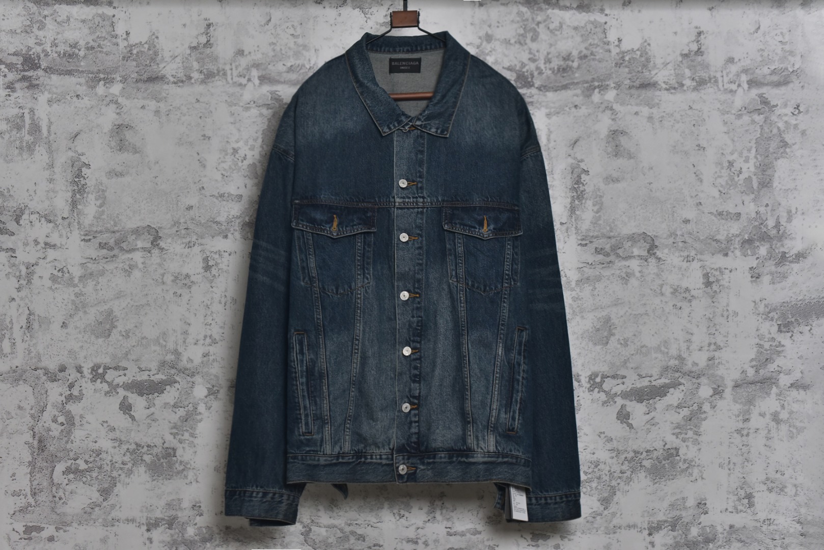 #Balenciaga/巴黎世家 25Fw 银胶带印花蓝牛仔外套 定制赛络纺工艺牛仔面料。此工艺使两根粗纱在纺纱阶段保持一定间距，经牵伸后由前罗拉输出，再汇合加捻成一根单纱。 赛络纺技术使最终织造出的丹宁面料具有卓越的强度、优异的耐磨性以及独特的柔软手感。纱线结构紧密，面料纹理清晰、光泽柔和，布面毛羽显著减少，呈现出普通牛仔无法比拟的细腻、挺括且高质感的视觉和触觉体验，为后续的重工艺处理提供了完美的基础。所有接缝、拼接处均采用高张力、高密度、均匀一致的针距进行缝制。线迹精准如机械制图，无任何跳针、断线或线头。内侧包边处理工整，承受高强度穿着与洗涤后仍能保持结构稳定，体现了世界级大厂对生产流程的严苛品控和无可挑剔的缝制技术。印花工艺复杂性： 这是整件外套最具技术挑战性的部分，其“胶带”效果并非普通印花或贴膜，而是采用了极为繁琐的多层厚板胶浆印花与特种闪粉混合印刷技术。厚板印刷： 通过多次重复印刷特调高分子胶浆，在面料表面堆叠出具有显著3D立体浮雕感的“胶带”条纹。每一次印刷都需经过精确对位和高温烘干定型，确保图案边缘锐利如刀刻，厚度均匀一致，手感坚硬且极具冲击力。闪粉融合： 在胶浆未完全固化前，将定制的高折射率微闪片通过精密筛网均匀撒在印花表面，并使其牢固嵌入胶浆内部。此步骤需精确控制胶浆粘度和闪粉的附着力，确保闪粉分布均匀、牢固不脱落，且光泽璀璨持久。 品质体现： 最终形成的印花图案不仅具有强烈的视觉立体感和触摸感，更因其高强度的物化特性，实现了极高的耐撕裂性、抗皱性和耐洗牢度。印花部分不会开裂、剥落或变黄，闪粉在日常穿着中也能最大限度地保持其闪耀效果，经久如新。这种工艺将装饰性与功能性推向了极致，成本高昂，良品率极低，是大厂才能驾驭的复杂技术 尺码：1 2 3-选品中心