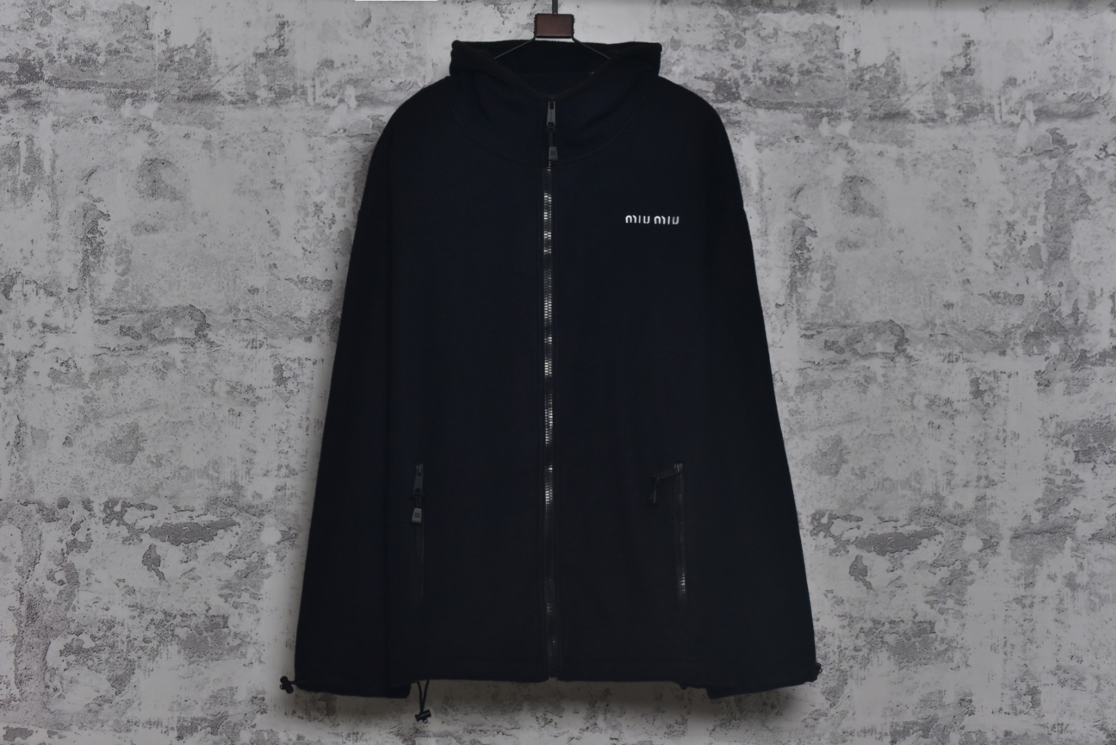 #Miu Miu 25Fw Miu小标摇粒绒连帽外套 面料采用定制织造染整一体工艺，从纱线选配到染色均按品牌标准定制。400克重摇粒绒需平衡蓬松度与垂感，通过经纬密优化确保厚度一致且抗风保暖。色牢度控制：定染环节采用环保活性染料，经5次水洗测试确保色彩零偏差，实现桃粉、薄荷绿等高饱和度色调的纯净度。肌理处理：双刷双摇工艺打造绒毛丰盈且定向倒伏的立体质感，避免传统摇粒绒的臃肿感。全件拉链及配件均为开模定制，非市场公版零件。拉链齿工艺：采用哑光涂层YKK EXCELLA系列，顺滑度与耐久度超越工业标准，拉头内部结构加入消音硅脂。挂绳挂件设计：拉链片与挂绳采用超声波切割无缝拼接，尾端缀以激光雕刻Miu Logo的锌合金吊坠，单挂件经历12道冲压打磨工序。装饰刺绣均采用进口田岛多针头绣花机完成。3D立体绣法：先以平绣打底，再叠加链目绣与毛巾绣混合工艺，使图案凸起高度达1.2mm，触感柔软不硌肤。本品以工艺冗余度换取极致体验，从纱线定制到终端装配均执行高精度标准，将功能性细节转化为隐性奢华，重构时尚单品的价值维度 尺码：S-L-选品中心