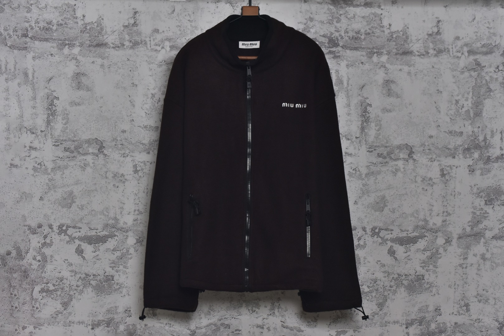 #Miu Miu 25Fw Miu小标咖色摇粒绒立领外套 面料采用定制织造染整一体工艺，从纱线选配到染色均按品牌标准定制。400克重摇粒绒需平衡蓬松度与垂感，通过经纬密优化确保厚度一致且抗风保暖。色牢度控制：定染环节采用环保活性染料，经5次水洗测试确保色彩零偏差，实现桃粉、薄荷绿等高饱和度色调的纯净度。肌理处理：双刷双摇工艺打造绒毛丰盈且定向倒伏的立体质感，避免传统摇粒绒的臃肿感。全件拉链及配件均为开模定制，非市场公版零件。拉链齿工艺：采用哑光涂层YKK EXCELLA系列，顺滑度与耐久度超越工业标准，拉头内部结构加入消音硅脂。挂绳挂件设计：拉链片与挂绳采用超声波切割无缝拼接，尾端缀以激光雕刻Logo的锌合金吊坠，单挂件经历12道冲压打磨工序。装饰刺绣均采用田岛多针头绣花机完成。3D立体绣法：先以平绣打底，再叠加链目绣与毛巾绣混合工艺，使图案凸起高度达1.2mm，触感柔软不硌肤。本品以工艺冗余度换取极致体验，从纱线定制到终端装配均执行高精度标准，将功能性细节转化为隐性奢华，重构时尚单品的价值维度 尺码：S-L-选品中心