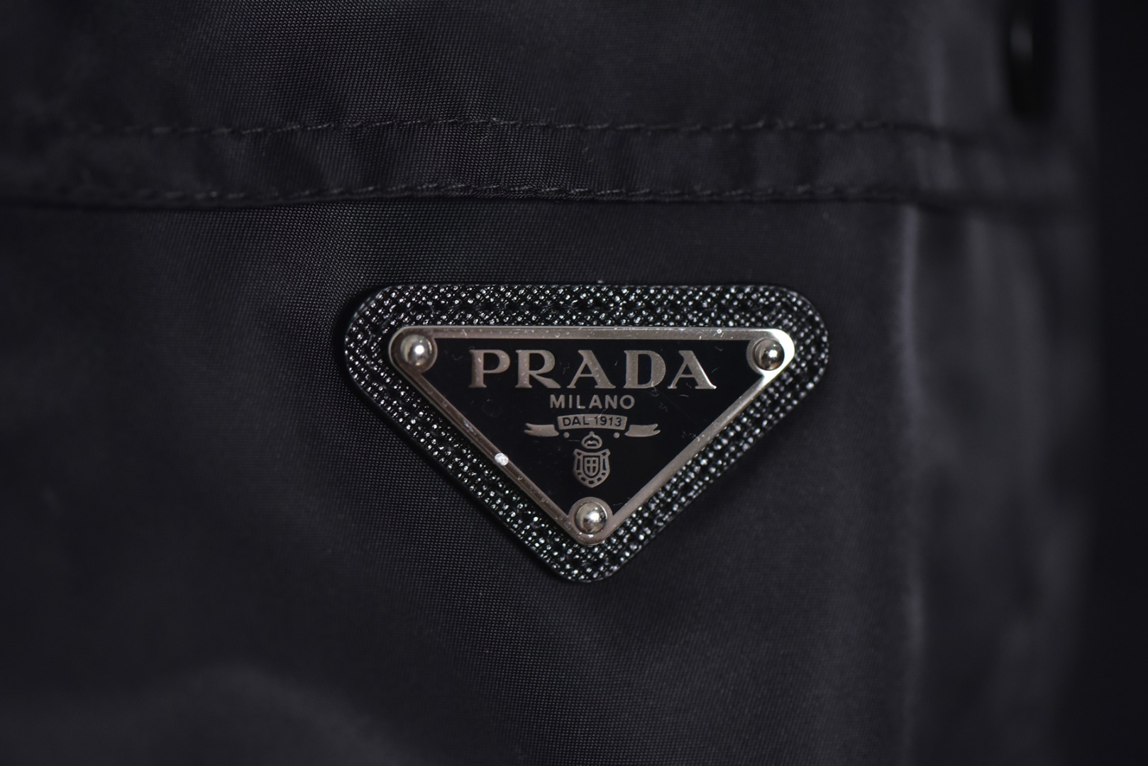 图片[3]-#Prada/普拉达 25Fw 双口袋三角标夹克外套 面料为特殊定制的高密度再生尼龙，并非市场普通现货，其密度、手感与防泼水性能均对标原版，力求还原Re-Nylon材质的轻盈质感与耐磨特性，原版面料重量精密控制在600-800克之间​（视款式而定），本品同样追求这种轻盈与挺括的平衡。所有五金件（包括拉链、扣具等）均源自东莞专业的箱包五金供应商，绝非普通市场通货，五金质感顺滑，经久耐用，拉链头顺滑度优异，减少卡顿感，与原版追求的耐用性一脉相承。胸前标志性的涂珐琅三角形金属徽标，采用的是与原厂（Prada供应商）一致的KF镜面三角标。此徽标由价值千万元的专用机器制作，呈现出镜面效果与清晰字体，细节精度远超市场仿品。原版徽标同样注重质感，其珐琅涂层通过盐雾测试，长期使用不易氧化。三标与洗标均为按原版材质定制，手感与材质均力求一致，确保细节处的正宗感。原版夹克在缝线工艺上采用双层缝线，拼接处使用隐藏式针脚，以提升耐用性和美观性，男女同款 尺码：46 48 50 52-选品中心