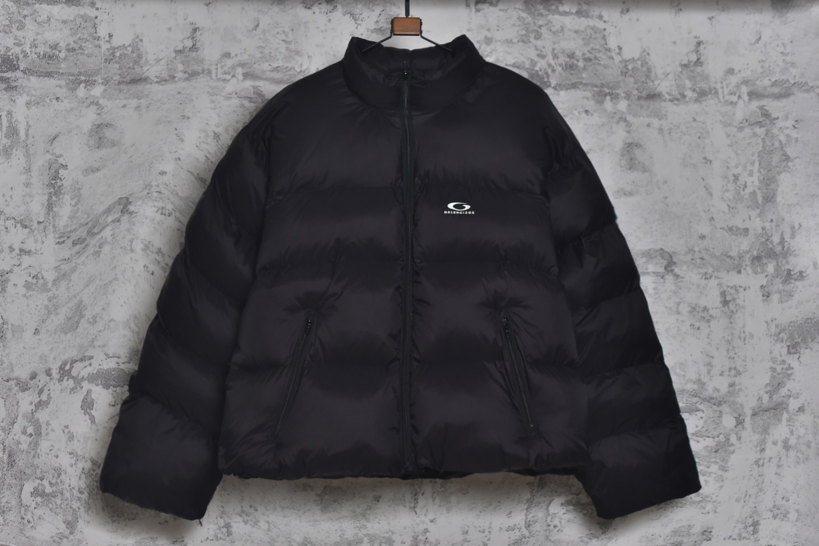 #Balenciaga/巴黎世家 25Fw 圆环小标棉服外套 定制开发高支高密梭织面料，是其高级质感的基石，高支数意味着纱线极细，从而织出纹理细腻、触感丝滑的表面，高密度则赋予了面料出色的挺括度、耐磨性与抗风能力，它避免了普通棉帆布的粗糙感，呈现出一种低调而精准的微光泽，为后续的刺绣与包边工艺提供了完美且稳定的基础，填充物选用珍珠棉，这是一种中空三维卷曲纤维，相比传统絮片，珍珠棉能提供更持久、均匀的蓬松感与回弹性，确保轮廓饱满不塌陷，中空结构可以锁住空气，实现最佳保暖重量比，达成“轻盈如羽，温暖如裘”的穿着体验，填充工艺需确保棉体在绗缝格内均匀分布，无空洞或结块，这要求极高的充绒精度。在此单品中，所有裁片边缘均采用斜裁包边条进行全方位包裹。这并非简单的锁边。高密度针迹，采用数万针的高密度刺绣，图案饱满立体，边缘清晰锐利，无任何松散线头，底层面料稳定处理，在如此轻薄高密的面料上进行密集刺绣，极易导致面料收缩变形，会在刺绣底层使用特制水溶衬或热熔衬，刺绣完成后这些衬布会消失，从而确保图案平整挺括，且不破坏面料本身的柔韧度。这是对手工与科技结合的极致考验，定制辅料三标，男女同款 尺码：XS-M-选品中心