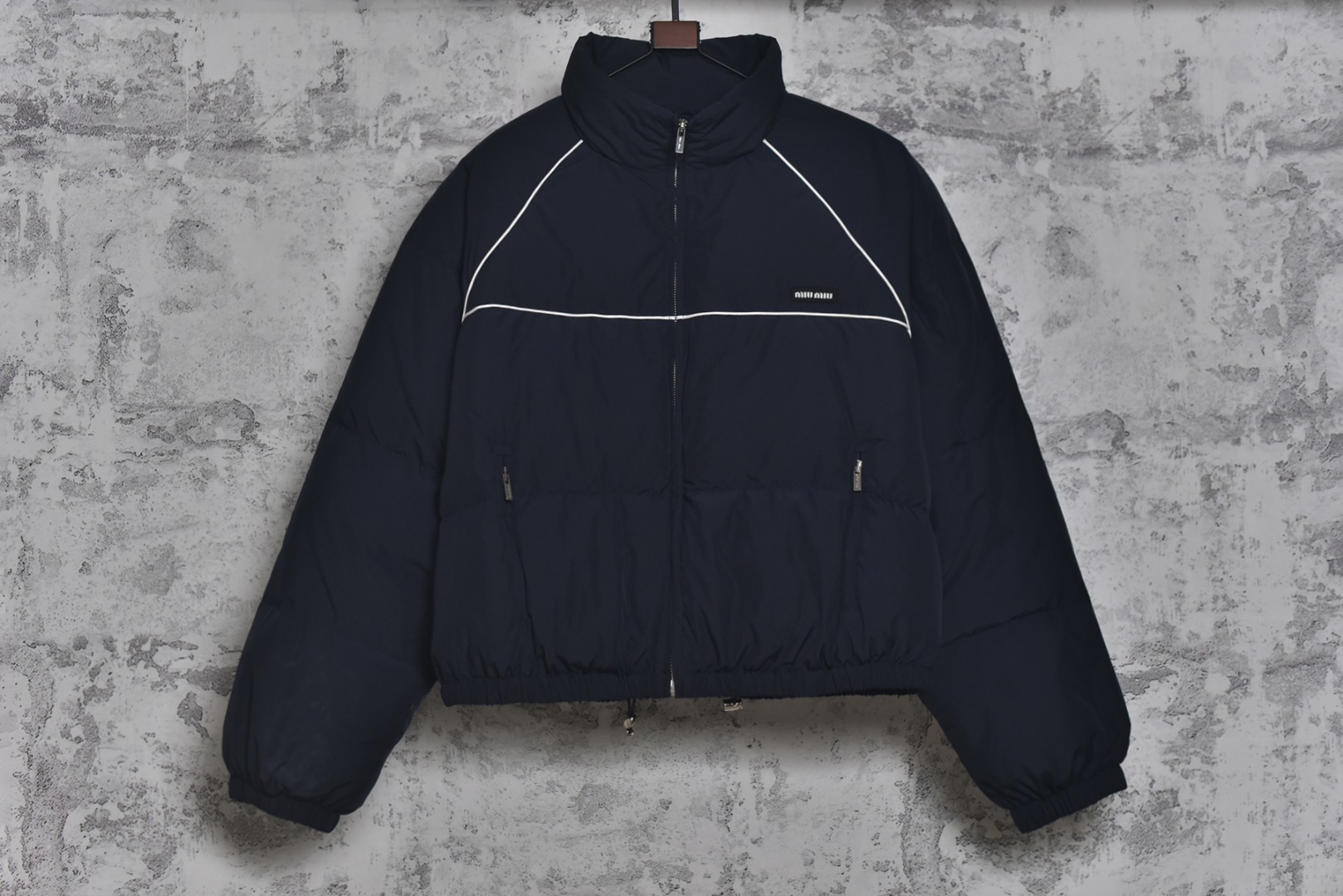 #Miu Miu 25Fw Miu小标捆条短款羽绒服 -核心内里填充180G广检认证90%纯白鸭绒 定制开发高支高密梭织面料，其纱线结构与织法经过精密计算，旨在实现极佳的防绒钻性，同时保证面料本身的轻盈与耐久度，面料色彩为独家定染，确保了色彩的纯粹度与独特性，采用捆条拼接工艺，该工艺在每块裁片的接缝处内衬一条白色织带，再将面料拼接并压线固定，白色的捆条在深色面料上形成了鲜明而利落的轮廓线，强化了单品的结构化视觉，内里填充90白鸭绒，高纯度绒子能锁住大量空气，形成静止空气层，实现高效保暖，这使得羽绒服在拥有极强保暖性的同时，重量得以大幅减轻，选用进口电脑充绒设备，确保每一个独立绗格内的羽绒填充量都精准、均匀，避免出现局部空洞或结块，从而保证羽绒服整体的平整度、保暖均匀性与持久蓬松，定制辅料三标 尺码：38 40 42-选品中心