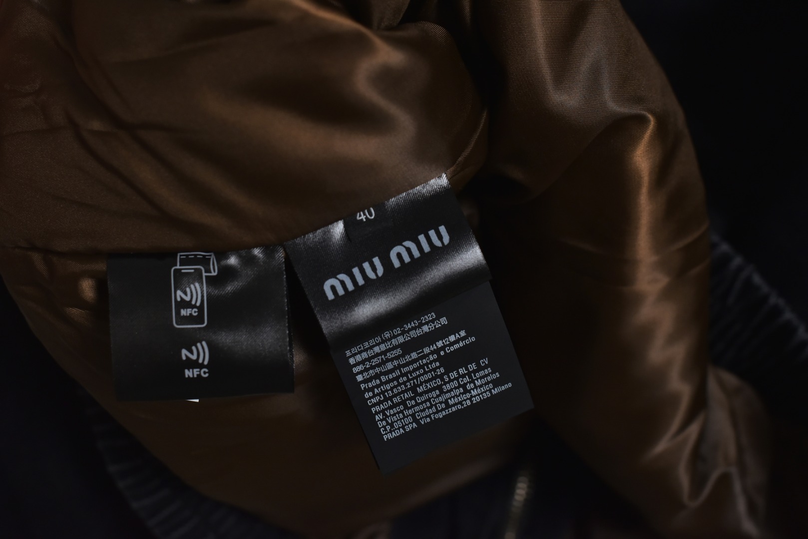 图片[9]-#Miu Miu 25Fw Miu小标牛仔棉服外套 选用高克重100%棉制牛仔斜纹布，高克重意味着面料单位面积密度更高，这不仅赋予了服装卓越的耐用性与抗皱性，更关键的是，它能自然支撑起利落而硬挺的立体廓形，无需依赖额外的内衬，面料经过专业的洗水处理，通过精准控制的酶洗、石磨等工艺，软化了原生棉布的僵硬感，使其触感更为亲肤柔顺。同时，这一过程赋予了丹宁独特的落色效果与微妙的复古做旧纹理，领部采用100%纯棉粗棱灯芯绒，其独特的瓦楞状脊线纹理与主体丹宁的斜纹肌理形成强烈而巧妙的视觉与触觉对比，将柔软且结构相对松散的灯芯绒与硬挺的牛仔布进行拼接，需要极高的裁切与缝纫精度，以确保接缝处平整牢固，避免因材质缩率不同而产生的变形。此举体现了品牌对异材质拼接工艺的成熟掌控，内里填充1400D高密度洗水棉。这里的“D”并非填充重量，而是指棉絮本身的纤维细度与长度规格，其拥有极佳的蓬松度与持久性，不易结块或移位，能确保夹克长期使用后仍保持均匀平整的保暖层，结合高克重牛仔布形成的“外壳结构”，共同构建了一个轻量却高效的隔热系统，实现了保暖性与廓形感的统一，在左前幅，运用了极为精细的真皮激光镭射雕花工艺。首先将一块小牛皮复合于面料之上，随后通过高精度激光按照预设图案进行烧蚀，刻画出品牌标识或图案，此工艺要求对激光的功率与速度进行毫厘不差的控制，才能在皮革上实现边缘锐利、细节分明的图案，同时不伤及底层的丹宁面料，定制辅料三标，男女同款 尺码：40 42 44-选品中心