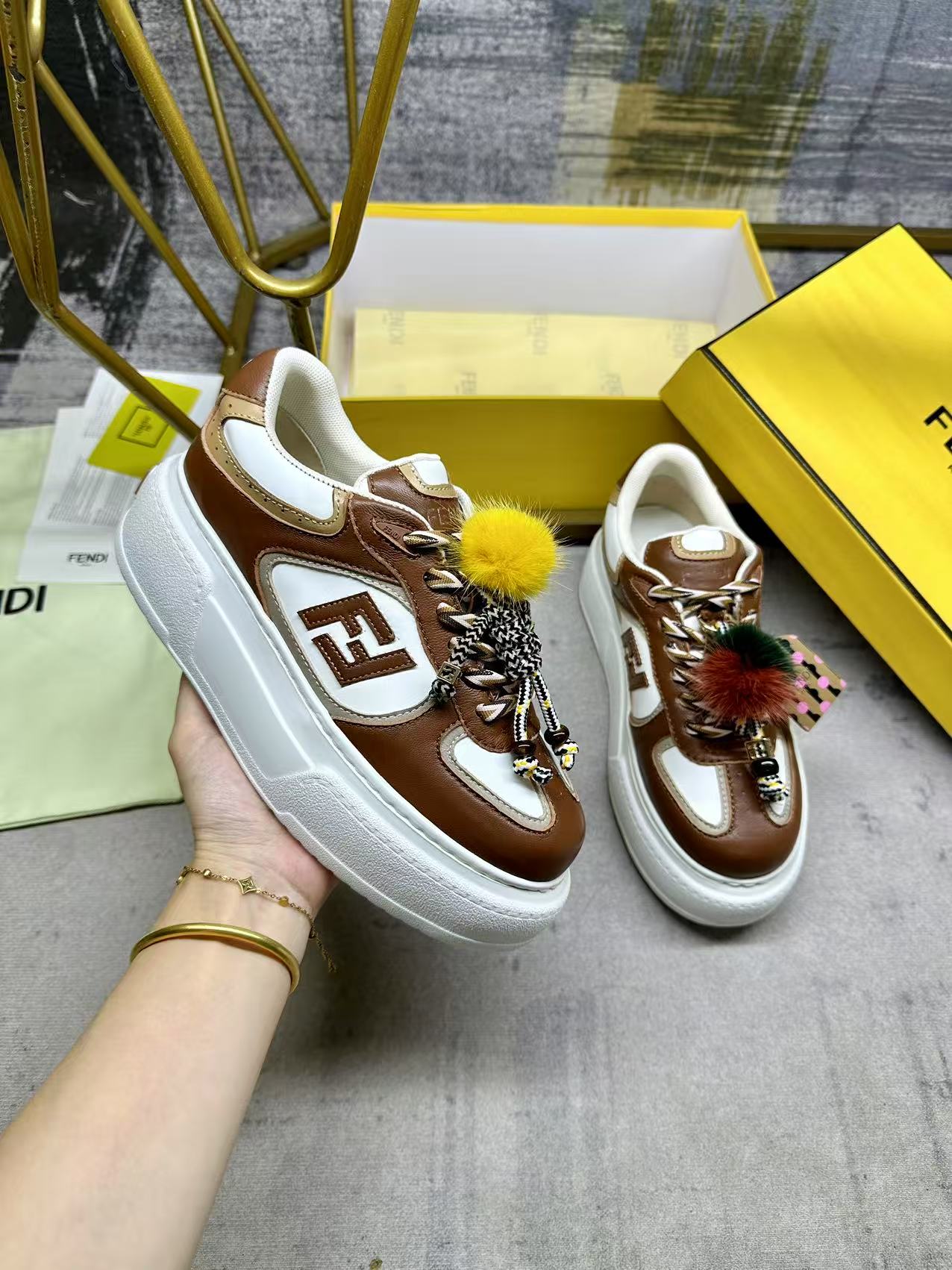 NO:592484,FENDI  Match lace-up sneakers. Brown fabric with beige details. Contrast leather FF appliqués on the sides and hiking laces in white, beige and brown. Thick rubber sole with oversized raised embossed FENDI lettering at the heel. Metal ring charm on the tongue. Contrast mink detailing and gold metallic F on the right shoe.  Small braided doll with F pattern bracelet. The left shoe is decorated with a braided rope with contrasting mink double pom-poms, decorated with metal FF beads and Pequin print pattern and logo (the doll is removable), sizes 35-42, casual shoes/sneakers, fendi, sneakers, bracelet19860909FENDI Match系带运动鞋.棕色面料,带米色细节.侧边饰有撞色皮革FF贴花,白色、米色和棕色登山鞋带.橡胶厚鞋底,后跟处饰有超大凸纹压花FENDI字样.鞋舌上饰有金属环挂饰右鞋上饰有带撞色貂毛细节和金色金属FF图案手链的编绳小玩偶.左鞋上饰有一条带撞色貂毛双绒球的编绳,饰有金属FF圆珠以及Pequin印花图案和标志（玩偶可拆卸）,35-42码,,休闲鞋/运动鞋,fendi,sneakers,bracelet,Women's Shoes