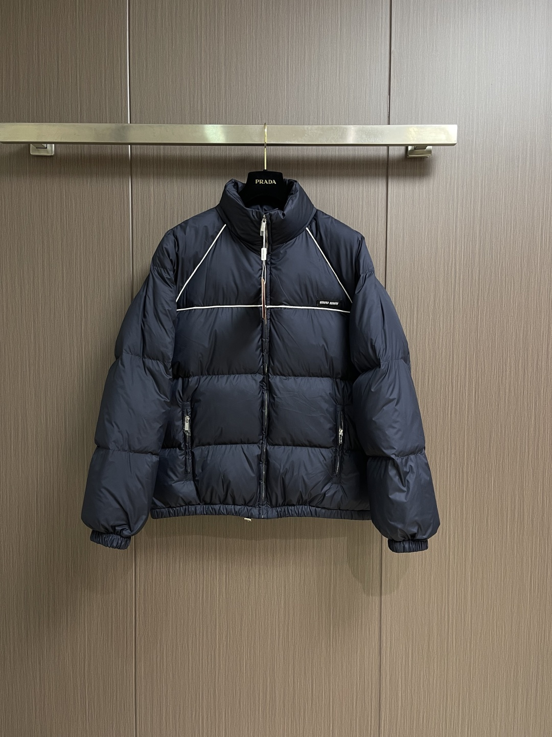 NO:600210,Miu  Miu patch-embroidered down jacket is made of nylon as the base material and uses a special weaving process to give the fabric surface a delicate texture, which feels as smooth as velvet but stiffer than traditional nylon. The fabric density is extremely high and the yarns are tightly arranged to form a natural windproof barrier while retaining moderate breathability to avoid stuffiness. As a down jacket outer  The layer of fabric can not only lock in the bulkiness of down, but also regulate body temperature through a breathable structure, maintaining a comfortable wearing experience in colder weather environments. It is filled with 90 white duck down, which is full and even, weighing 210g, ensuring good thermal insulation performance. The chest is embellished with a contrasting knitted logo patch, and the brand logo is outlined with precision embroidery.  The logo, white letter stitching and navy blue fabric are in sharp contrast, showing a sense of high-end sportiness in simplicity. The edges of the logo are hand-wrapped, and the delicate touch echoes the fabric texture subtly. The high-neck design fits the neck line, and is covered with delicate lining, which is windproof and cold-proof, without losing the silhouette, and shows a low-key sense of strength in every gesture. Pull  The chain head is embellished with MIUMIU's signature metal ornaments, which hides the brand imprint in a low-key manner. The oblique zipper pockets on both sides have ergonomic opening angles, making it easy to access items while ensuring the safety of items. The inside of the pocket is designed with velvet to keep your hands warm in the cold winter. Sizes 46-52,,miumiu,jackets  & coats,down jacket,alexander wang19860909Miu Miu饰贴布绣羽绒服外套,以尼龙为基材,采用特殊织造工艺,使面料表面呈现细腻的纹理,触感柔滑如丝绒,却比传统尼龙更具挺括度.面料密度极高,纱线排列紧密,形成天然防风屏障,同时保留适度透气性,避免闷热感.作为羽绒服外层面料,既能锁住羽绒蓬松度,又能通过透气结构调节体温,在较寒冷天气环境下均能保持舒适穿着体验.采用90白鸭绒填充饱满且均匀,克重达210g,保证了良好的保暖性能.胸口处点缀撞色针织徽标贴饰,以精密刺绣工艺勾勒品牌标识,白色字母缝线与藏青色面料形成鲜明对比,简约中透出高级运动感.徽标边缘采用手工包边处理,细腻触感与面料肌理形成微妙呼应.高领设计贴合颈部线条,内覆细腻里衬,既防风御寒,又不失利落廓形,举手投足间流露低调力量感.拉链头缀以MIUMIU标志性金属饰件,低调中暗藏品牌印记.两侧斜插式拉链口袋,开口角度经过人体工学考量,取物便捷,同时保障物品安全,口袋内侧加绒设计,寒冬里亦可温暖双手.尺码46-52,,miumiu,jackets & coats,down jacket,alexander wang,Men's clothing