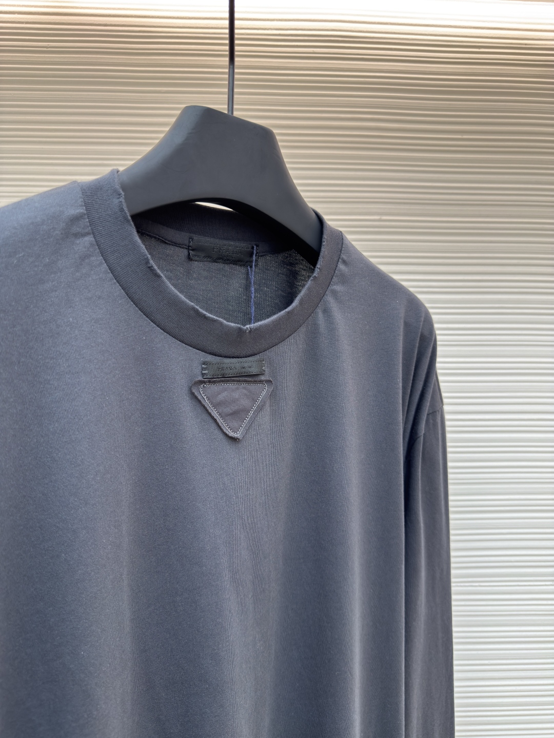 P125 Prada 铅笔灰长袖
●原版：8934购入开发
●货号:  1680720-A05
●面料：精梳平纹面料
●工艺：激光烧花制作
●版型：合身版型
●克重：约240克
●尺码：S-M-L-XL
●描述：定织50S 190G精梳平纹面料，非市场通用坯布，需从纱线配比、织机参数全程定制。通过精梳工艺去除短纤维杂质，确保纱线更长、更均匀，织物表面光洁无毛羽。  特殊定染铅笔灰色，单独调色而非使用标准色卡，需多次打样匹配Prada指定的低饱和度灰调。胸前三角激光烧花绣花定型：激光烧花：高精度激光雕刻，在面料表面碳化形成渐变蚀刻纹理，边缘需人工调试避免焦痕。  复合绣花：在烧花基础上叠加刺绣，线迹密度需与烧花图案对齐，防止面料变形。  立体定型：使用高温压烫或树脂涂层维持三角结构的浮雕感，避免洗后塌陷。整体走线平直细腻，面料舒适柔软，颜色一致无色差 能与同款搭配营造时尚造型，驾驭各种场合，定制辅料，三标齐全，男女同款！