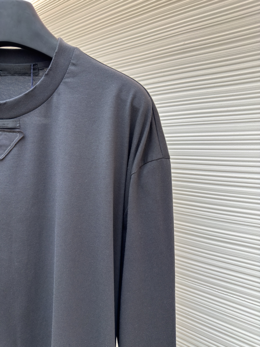 P125 Prada 铅笔灰长袖
●原版：8934购入开发
●货号:  1680720-A05
●面料：精梳平纹面料
●工艺：激光烧花制作
●版型：合身版型
●克重：约240克
●尺码：S-M-L-XL
●描述：定织50S 190G精梳平纹面料，非市场通用坯布，需从纱线配比、织机参数全程定制。通过精梳工艺去除短纤维杂质，确保纱线更长、更均匀，织物表面光洁无毛羽。  特殊定染铅笔灰色，单独调色而非使用标准色卡，需多次打样匹配Prada指定的低饱和度灰调。胸前三角激光烧花绣花定型：激光烧花：高精度激光雕刻，在面料表面碳化形成渐变蚀刻纹理，边缘需人工调试避免焦痕。  复合绣花：在烧花基础上叠加刺绣，线迹密度需与烧花图案对齐，防止面料变形。  立体定型：使用高温压烫或树脂涂层维持三角结构的浮雕感，避免洗后塌陷。整体走线平直细腻，面料舒适柔软，颜色一致无色差 能与同款搭配营造时尚造型，驾驭各种场合，定制辅料，三标齐全，男女同款！