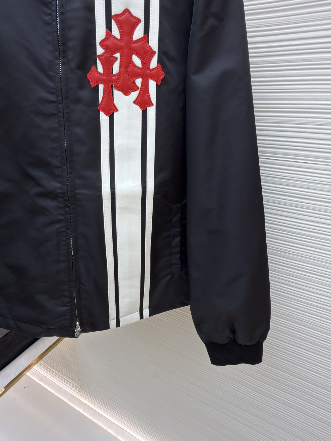 P295 Chrome Hearts 贴布条纹夹克
●原版：30000购入开发
●货号：1681765-B60
●面料：聚酯纤维面料
●工艺：贴布制作
●颜色：黑色
●版型：合身版型
●克重：约680克
●尺码：S-M-L-XL
●描述：采用高密防风面料，经防泼水涂层处理，兼具耐用性与防护性，触感顺滑且不易起皱。衣身侧边拼接黑白条纹装饰，通过精准定位缝制工艺固定，线条规整增强视觉层次；正面装饰品牌红色Logo印花，采用高温烫印工艺，色彩饱和且不易脱落，凸显辨识度。版型为宽松剪裁，搭配金属拉链开合，穿脱便捷；罗纹领口、袖口及下摆增强贴合度，兼顾防风性与休闲感。全衣缝线工整，拼接处无外露线头，手工测量尺寸（误差1-3CM属正常），工艺平衡了街头风格与品牌标志性设计，是兼具实穿性与潮流感的单品。整体走线平直细腻，面料舒适柔软，颜色一致无色差 能与同款搭配营造时尚造型，驾驭各种场合，定制辅料，三标齐全，男女同款！