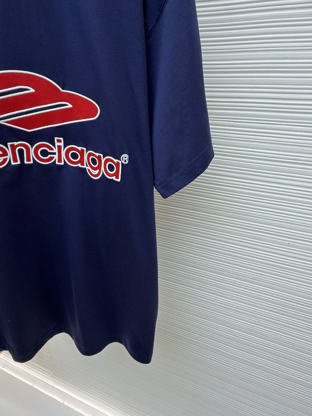 P180 Balenciaga 球队系列V领长袖
●原版：5699购入开发
●货号：1682041-A50
●面料：平纹针织紧密棉面料
●工艺：胶浆印花制作
●颜色：蓝色
●版型：宽松版型
●克重：约300克
●尺码：XS-S-M-L
●描述：主体采用24支特纺紧密棉，克重为185g/㎡。在纺纱环节应用了紧密纺技术，显著减少了纱线毛羽，使得纱线更光滑、强韧。这赋予了面料结构异常密实、表面光洁平整、抗起球性优异的特点，同时保留了纯棉的柔软透气性。185g的克重属于轻至中量级，确保了衣物轻盈、垂坠且活动自如的特性，非常适合打造兼具运动感与日常舒适度的长袖衫。V领部分由两种不同颜色的高弹罗纹面料拼接而成。其工艺步骤是先分别织造出两种颜色的罗纹带，再通过精准的剪裁与拼接，形成具有鲜明色彩对比的V形领口。打破了纯色衣身的单调，以强烈的色彩对比塑造出年轻、活力的视觉焦点，并优化了颈部线。将图案在不同材质或颜色的布料上预先裁剪成型贴布，然后将其覆盖在衣身上，最后用高密度绣线锁边固定并勾勒内部细节。其作用在于：创造色彩鲜明、质感对比强烈且极具立体感和耐用性的图案，图案边缘牢固，不易磨损。将特制的弹性浆料通过网版印刷在袖部的工艺，色彩鲜艳且具有轻微涂层感的图案。弹力浆料保证了印花区域能随面料一起拉伸而不开裂。与贴布绣的立体感形成一平一凸的质感对比，增加了设计的趣味性和复杂度。整体走线平直细腻，面料舒适柔软，颜色一致无色差 能与同款搭配营造时尚造型，驾驭各种场合，定制辅料，三标齐全，男女同款！