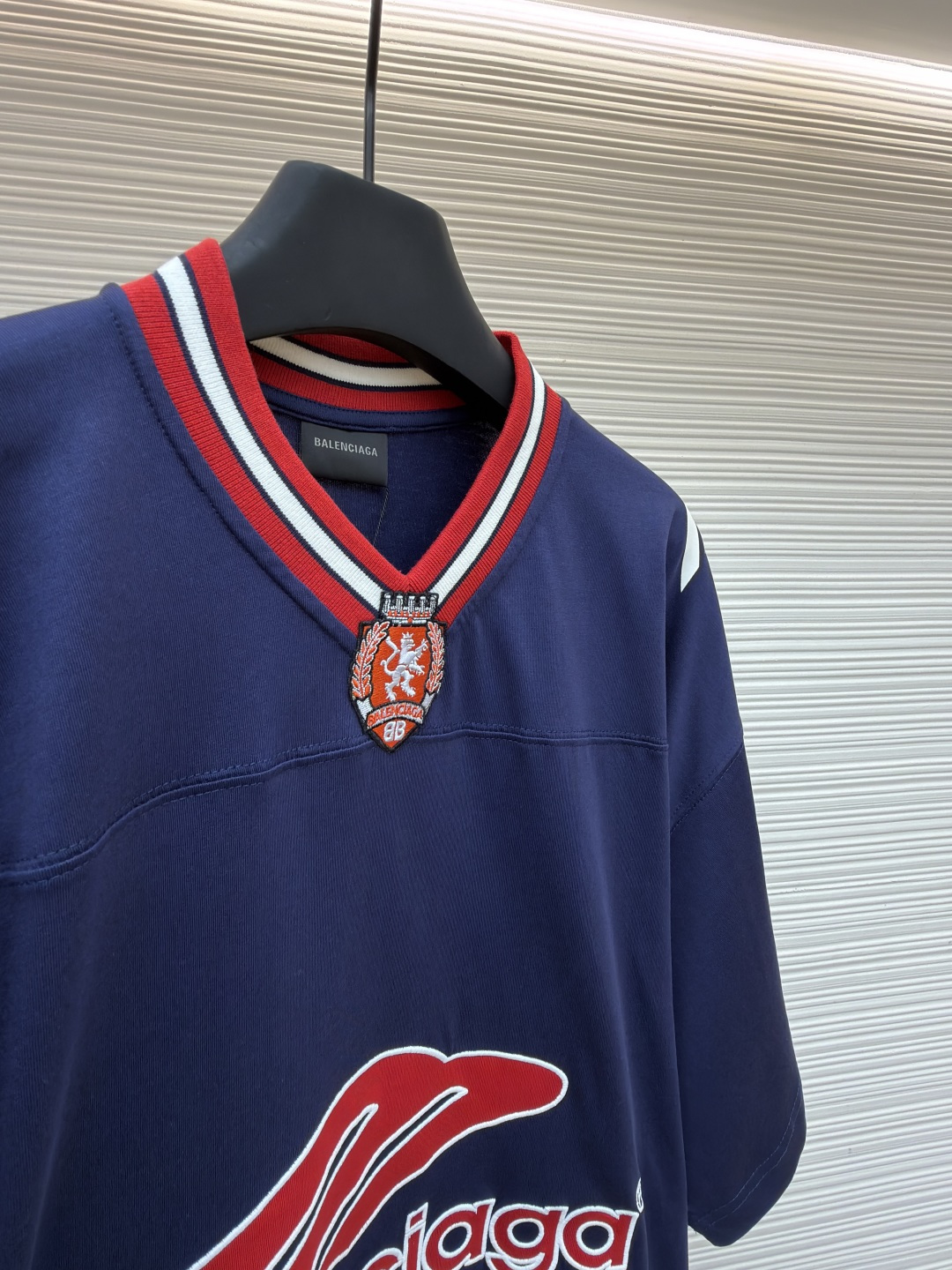 P180 Balenciaga 球队系列V领长袖
●原版：5699购入开发
●货号：1682041-A50
●面料：平纹针织紧密棉面料
●工艺：胶浆印花制作
●颜色：蓝色
●版型：宽松版型
●克重：约300克
●尺码：XS-S-M-L
●描述：主体采用24支特纺紧密棉，克重为185g/㎡。在纺纱环节应用了紧密纺技术，显著减少了纱线毛羽，使得纱线更光滑、强韧。这赋予了面料结构异常密实、表面光洁平整、抗起球性优异的特点，同时保留了纯棉的柔软透气性。185g的克重属于轻至中量级，确保了衣物轻盈、垂坠且活动自如的特性，非常适合打造兼具运动感与日常舒适度的长袖衫。V领部分由两种不同颜色的高弹罗纹面料拼接而成。其工艺步骤是先分别织造出两种颜色的罗纹带，再通过精准的剪裁与拼接，形成具有鲜明色彩对比的V形领口。打破了纯色衣身的单调，以强烈的色彩对比塑造出年轻、活力的视觉焦点，并优化了颈部线。将图案在不同材质或颜色的布料上预先裁剪成型贴布，然后将其覆盖在衣身上，最后用高密度绣线锁边固定并勾勒内部细节。其作用在于：创造色彩鲜明、质感对比强烈且极具立体感和耐用性的图案，图案边缘牢固，不易磨损。将特制的弹性浆料通过网版印刷在袖部的工艺，色彩鲜艳且具有轻微涂层感的图案。弹力浆料保证了印花区域能随面料一起拉伸而不开裂。与贴布绣的立体感形成一平一凸的质感对比，增加了设计的趣味性和复杂度。整体走线平直细腻，面料舒适柔软，颜色一致无色差 能与同款搭配营造时尚造型，驾驭各种场合，定制辅料，三标齐全，男女同款！