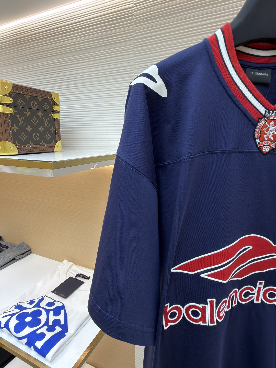 P180 Balenciaga 球队系列V领长袖
●原版：5699购入开发
●货号：1682041-A50
●面料：平纹针织紧密棉面料
●工艺：胶浆印花制作
●颜色：蓝色
●版型：宽松版型
●克重：约300克
●尺码：XS-S-M-L
●描述：主体采用24支特纺紧密棉，克重为185g/㎡。在纺纱环节应用了紧密纺技术，显著减少了纱线毛羽，使得纱线更光滑、强韧。这赋予了面料结构异常密实、表面光洁平整、抗起球性优异的特点，同时保留了纯棉的柔软透气性。185g的克重属于轻至中量级，确保了衣物轻盈、垂坠且活动自如的特性，非常适合打造兼具运动感与日常舒适度的长袖衫。V领部分由两种不同颜色的高弹罗纹面料拼接而成。其工艺步骤是先分别织造出两种颜色的罗纹带，再通过精准的剪裁与拼接，形成具有鲜明色彩对比的V形领口。打破了纯色衣身的单调，以强烈的色彩对比塑造出年轻、活力的视觉焦点，并优化了颈部线。将图案在不同材质或颜色的布料上预先裁剪成型贴布，然后将其覆盖在衣身上，最后用高密度绣线锁边固定并勾勒内部细节。其作用在于：创造色彩鲜明、质感对比强烈且极具立体感和耐用性的图案，图案边缘牢固，不易磨损。将特制的弹性浆料通过网版印刷在袖部的工艺，色彩鲜艳且具有轻微涂层感的图案。弹力浆料保证了印花区域能随面料一起拉伸而不开裂。与贴布绣的立体感形成一平一凸的质感对比，增加了设计的趣味性和复杂度。整体走线平直细腻，面料舒适柔软，颜色一致无色差 能与同款搭配营造时尚造型，驾驭各种场合，定制辅料，三标齐全，男女同款！